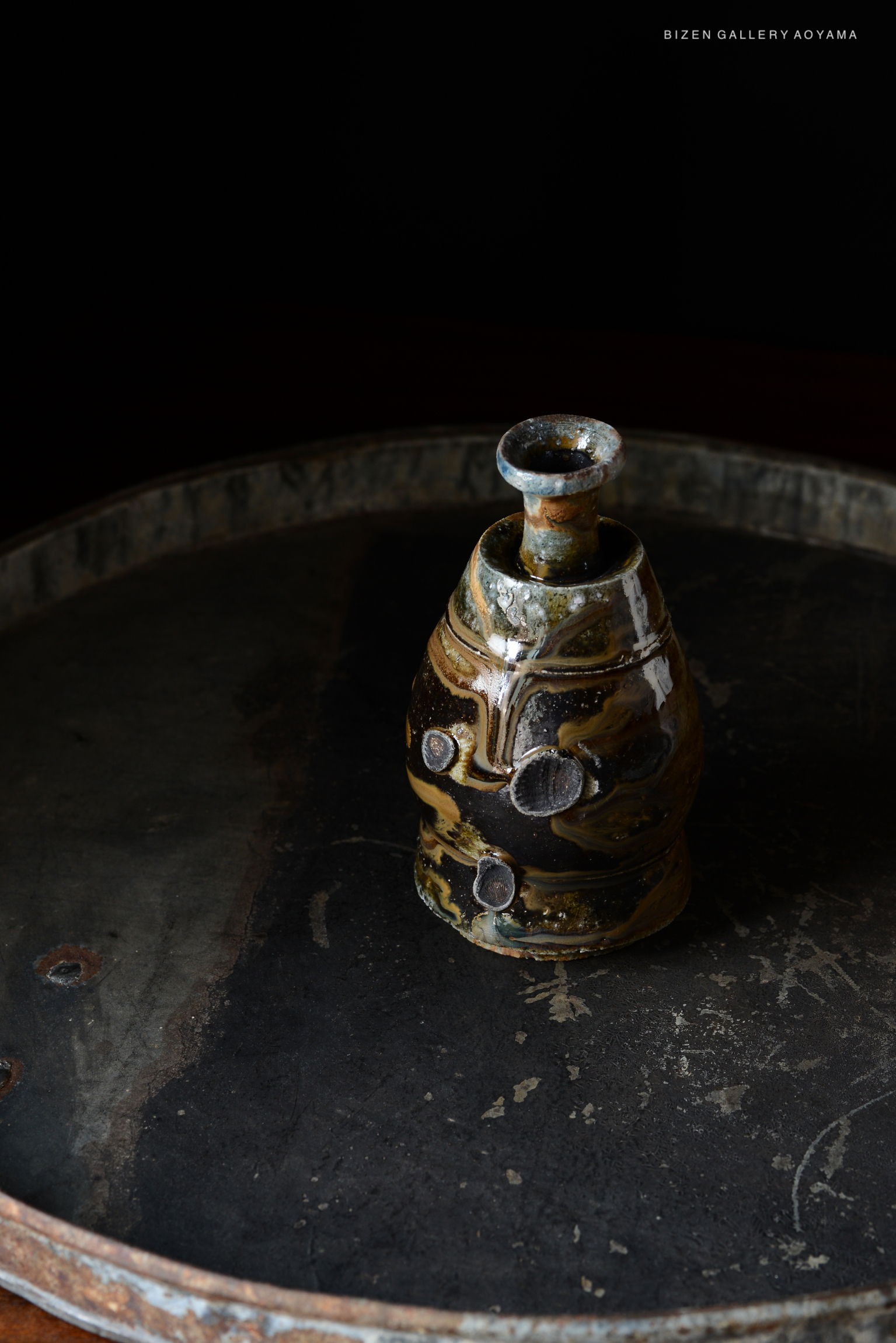 A traditional Kuro Tokkuri, a Japanese sake vessel, displayed on a textured circular surface with a dark background.