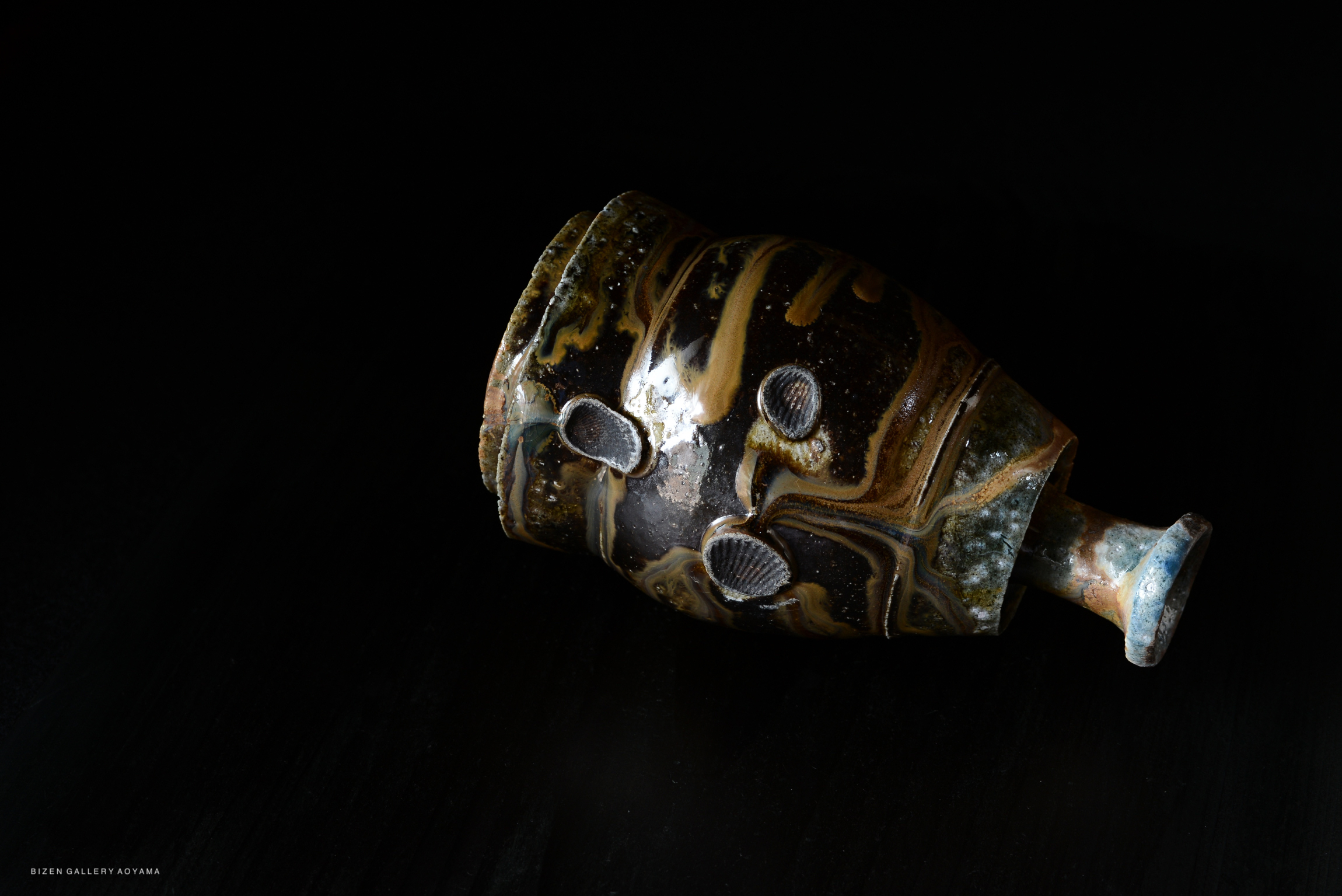 A close-up of a black and earthy-toned Japanese Tokkuri sake bottle, featuring unique textured patterns and a narrow spout, set against a dark background.