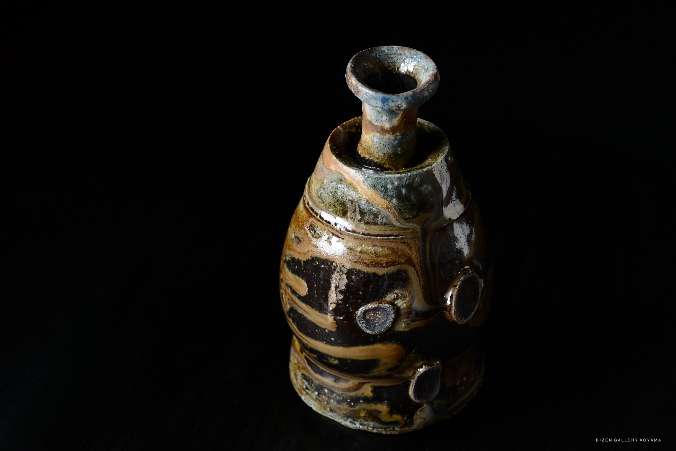 A handcrafted black and brown ceramic Tokkuri (sake vessel) with a unique glaze pattern, set against a dark background.