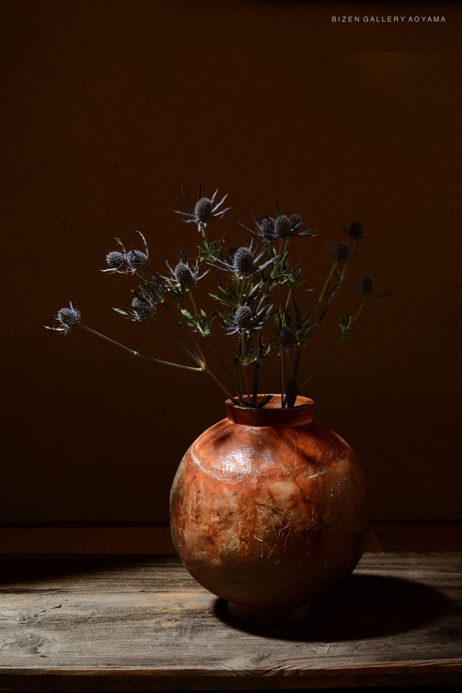 A Bizen vase with a round shape and cracked surface, holding dried thistle flowers, placed on a wooden table with a subtly lit background.