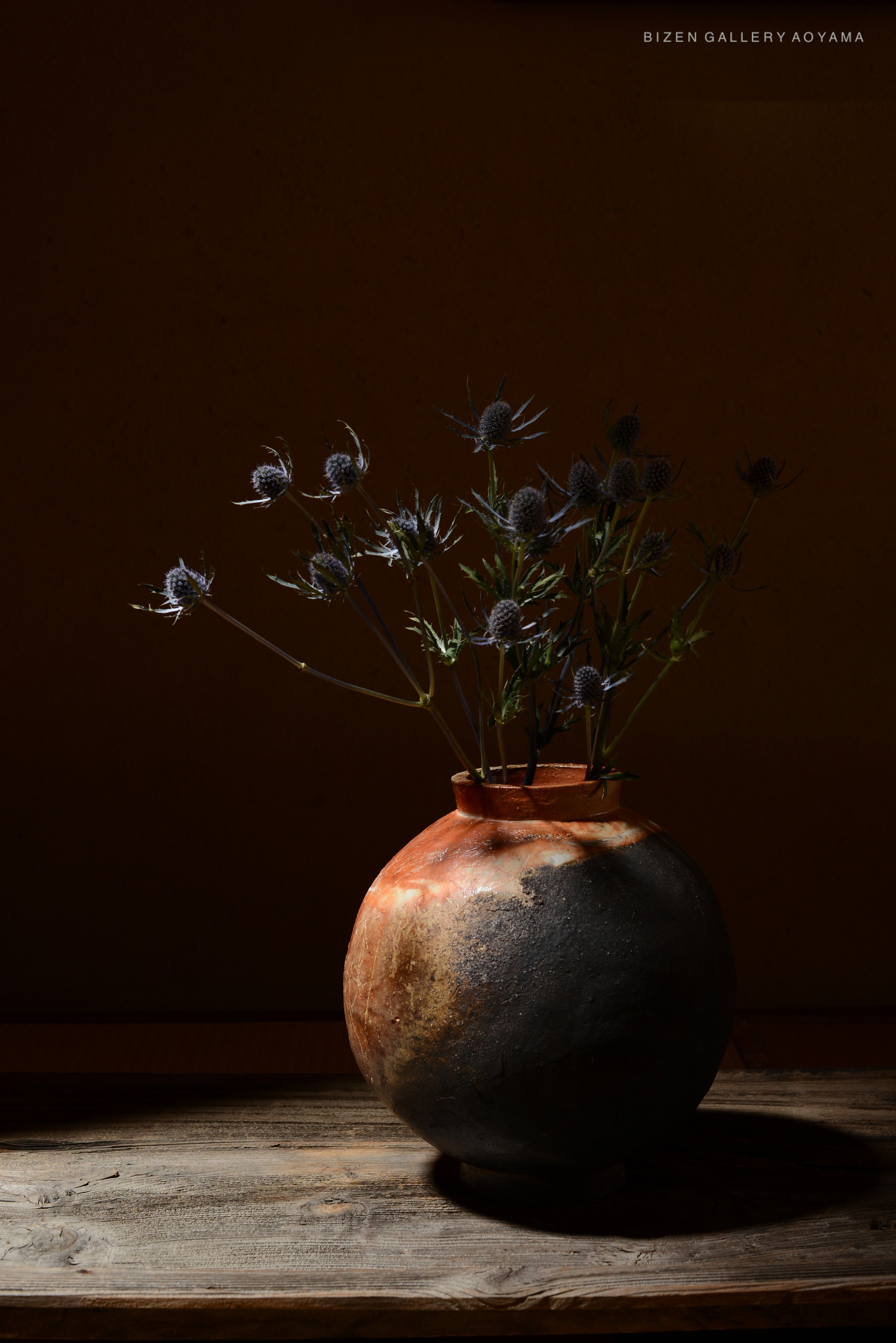 A rustic Bizen pottery vase with flowers, illuminated by soft natural light, placed on a wooden table against a warm, textured wall.