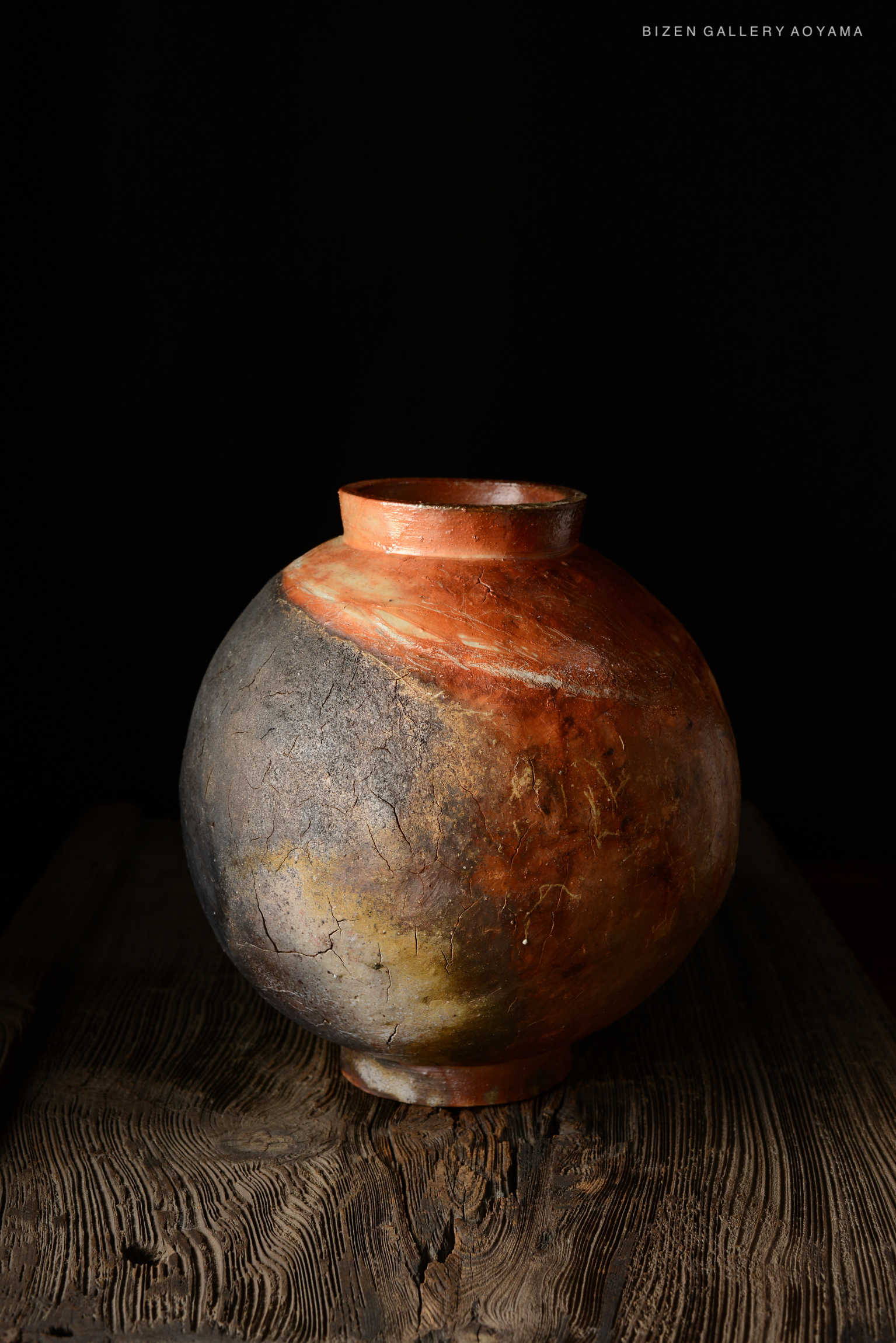 A handcrafted Bizen pottery vase with a smooth, rounded body and a narrow neck, featuring a blend of earthy colors in shades of orange, gray, and black, placed on a rustic wooden surface.