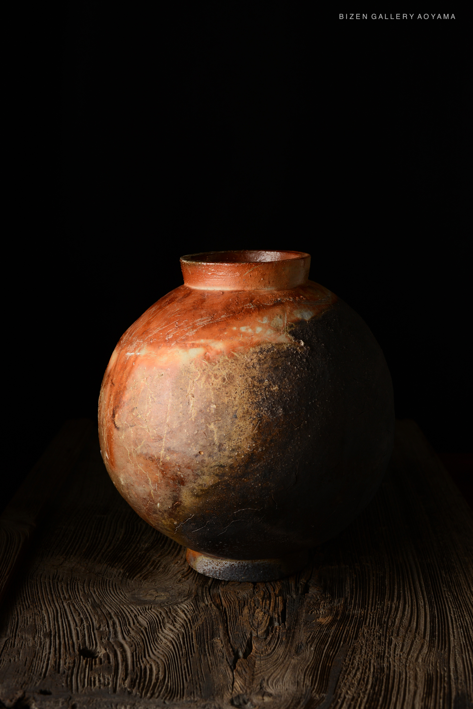 A round Bizen pottery vase with a textured surface, featuring earthy tones of orange and black, set against a dark background.