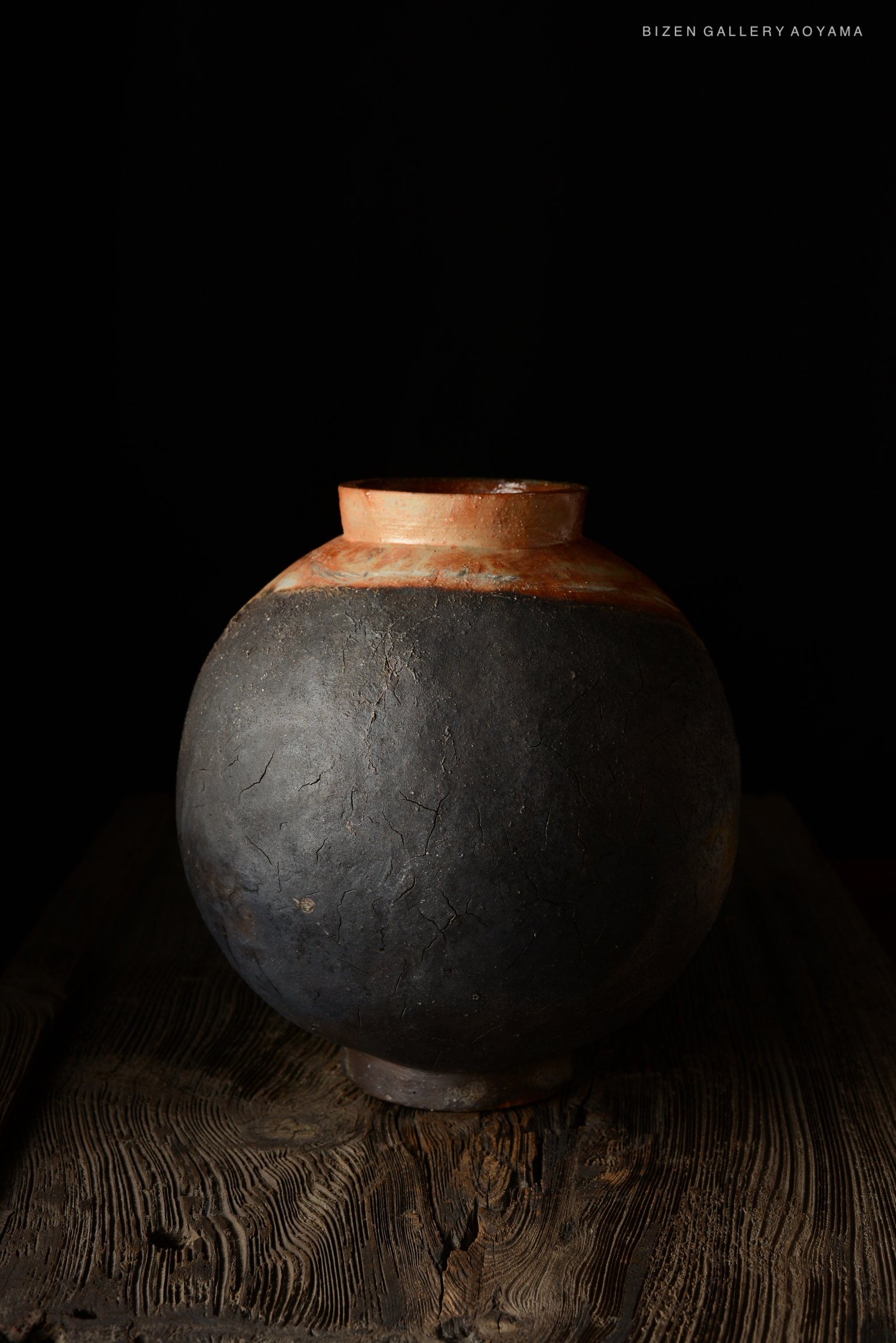 A traditional Bizen pottery jar with a rounded black body and a light-colored rim, placed on a wooden surface against a dark background.