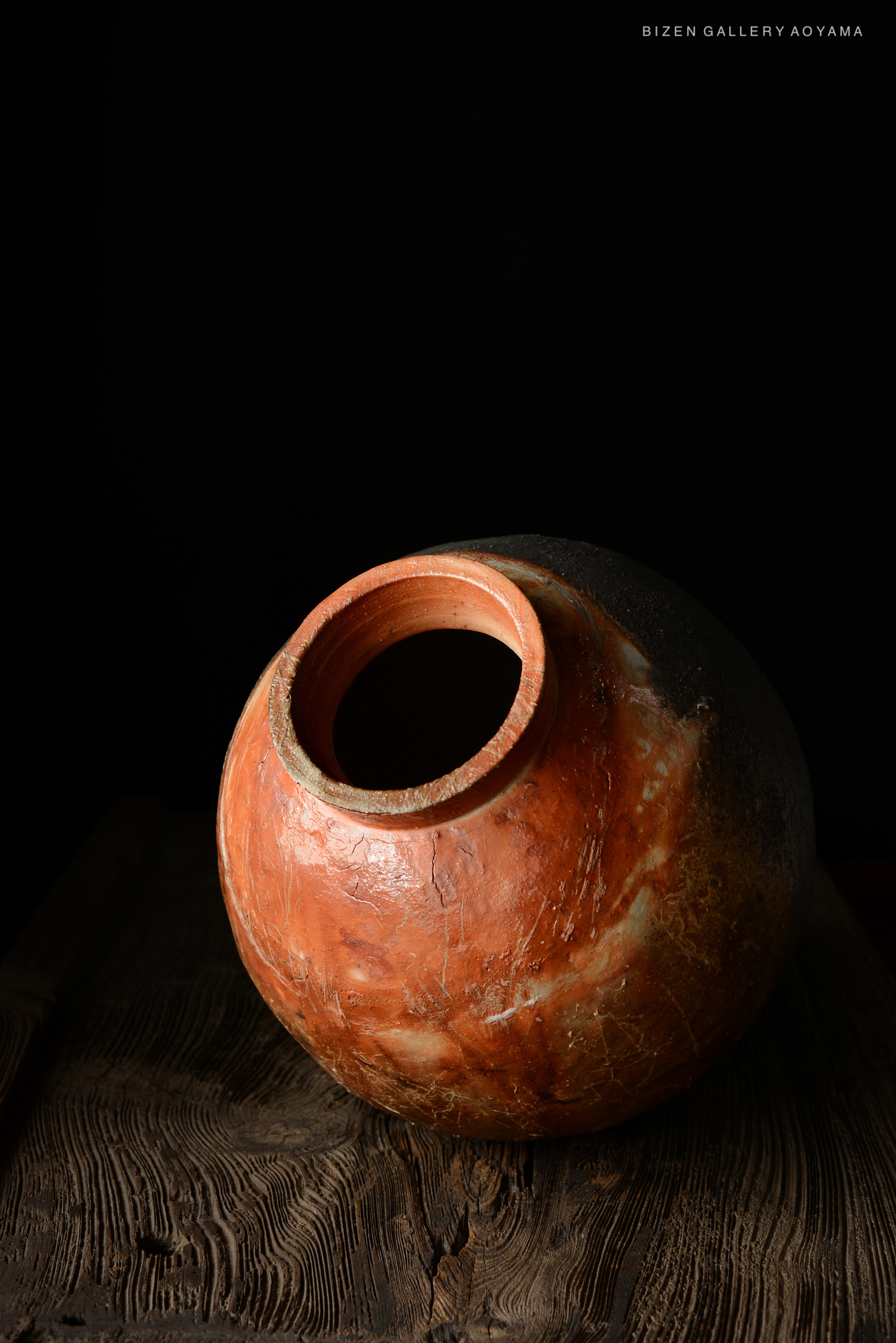 A handcrafted Bizen pottery vase with a rounded body and a narrow neck, sitting on a rustic wooden surface, against a black background.