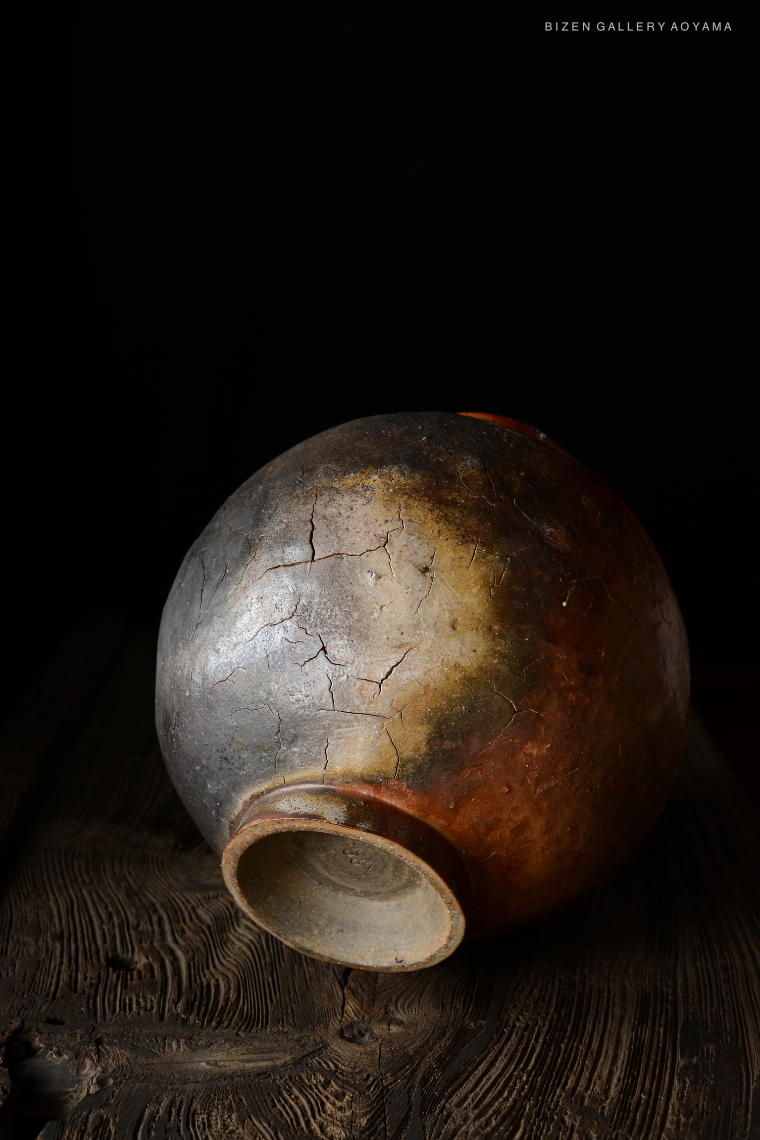 A close-up view of a cracked Bizen pottery vase with a smooth, rounded shape and a dark, weathered finish, resting on a wooden surface.