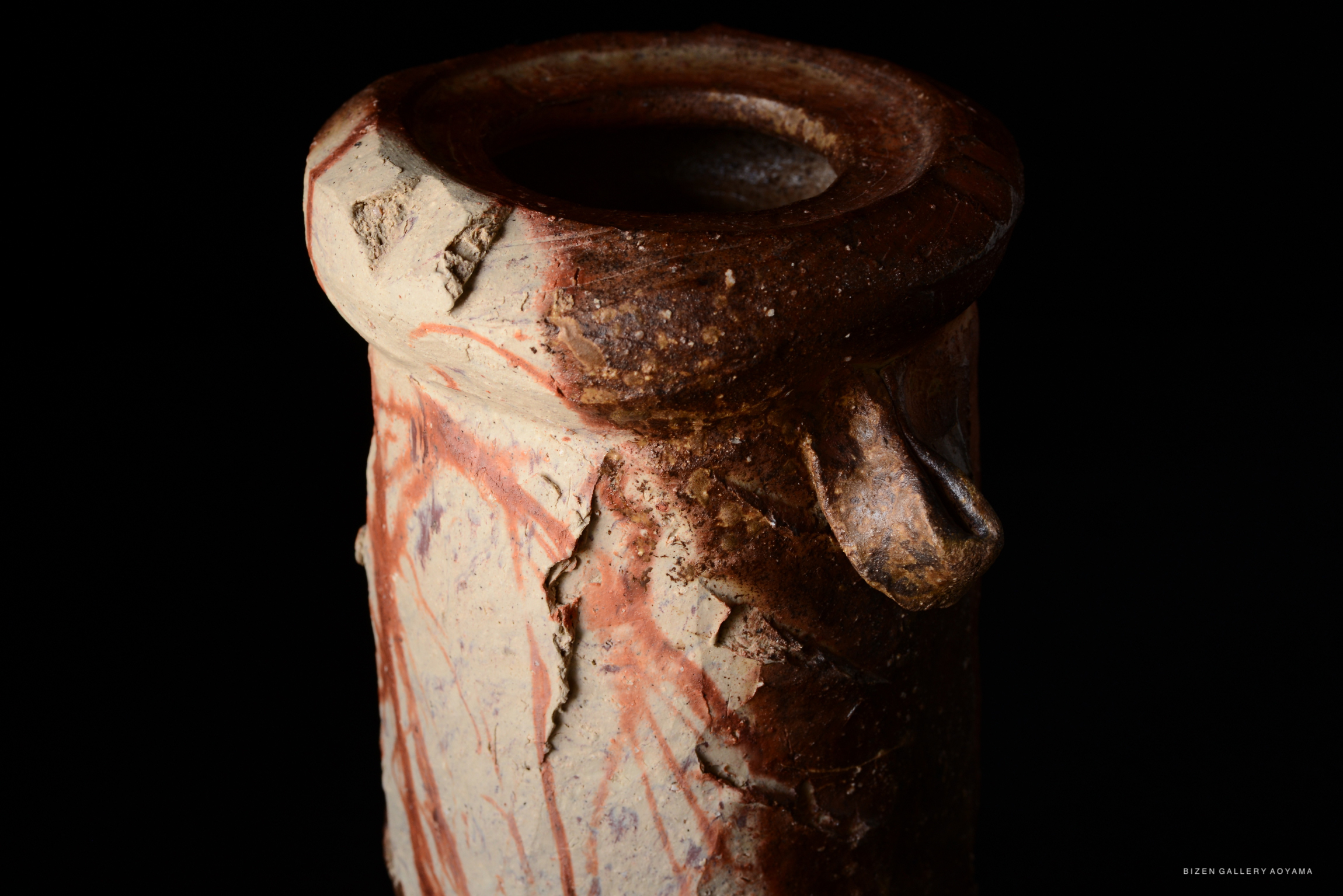 Close-up view of a ceramic tsubo with textured surface and reddish-brown streaks, set against a dark background.