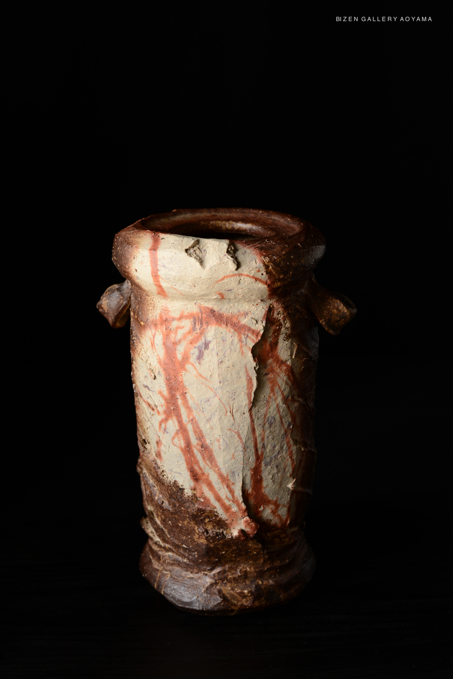 A ceramic vessel with a textured surface and natural coloring, featuring subtle red streaks and a robust base, displayed against a dark background.