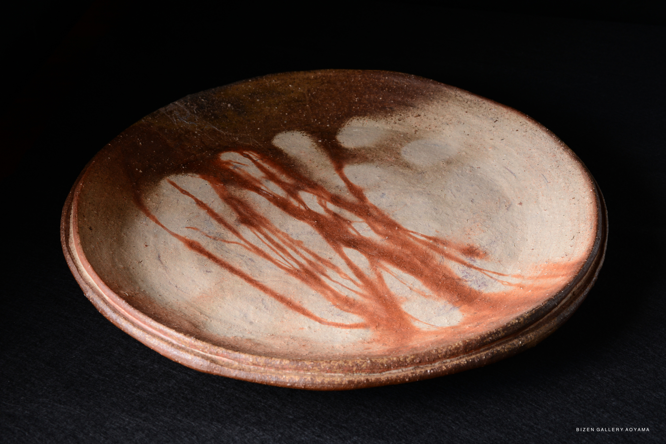 A large, shallow ceramic plate with a natural glaze, showcasing streaks of reddish-brown markings on a light clay surface.