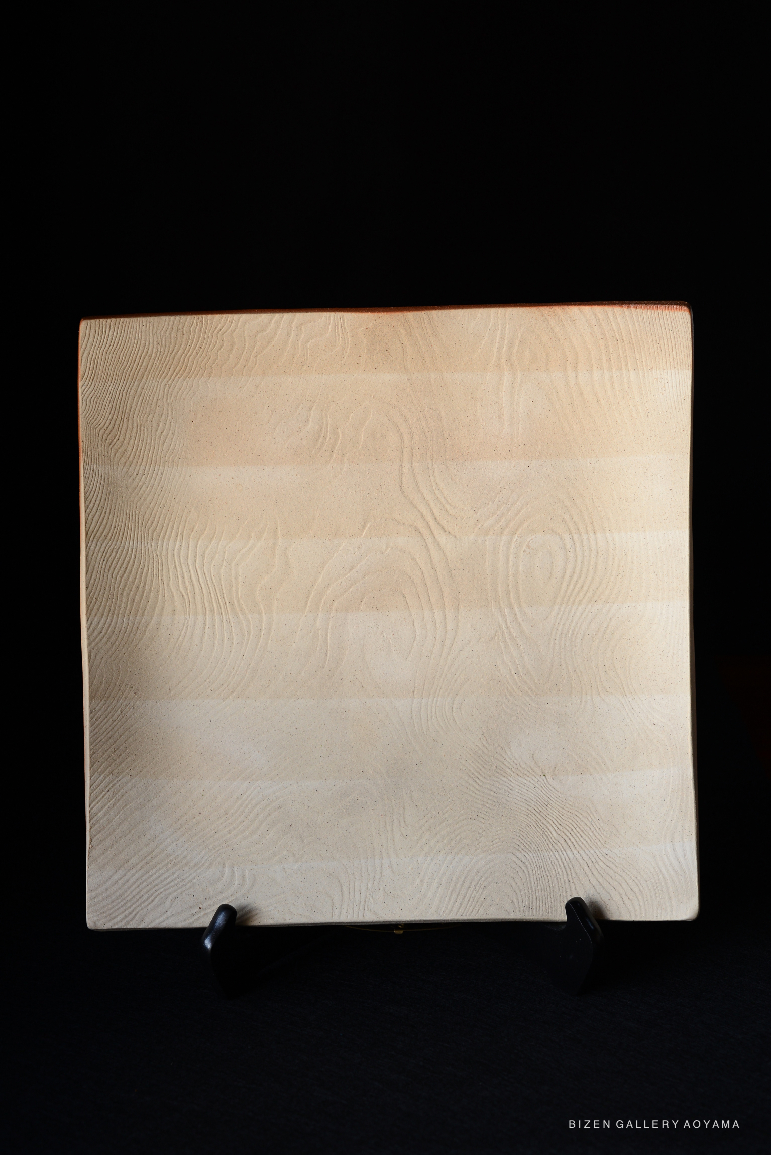 A simple, elegant ceramic plate with a smooth, textured surface, displaying natural wood-like patterns, presented on a black stand.
