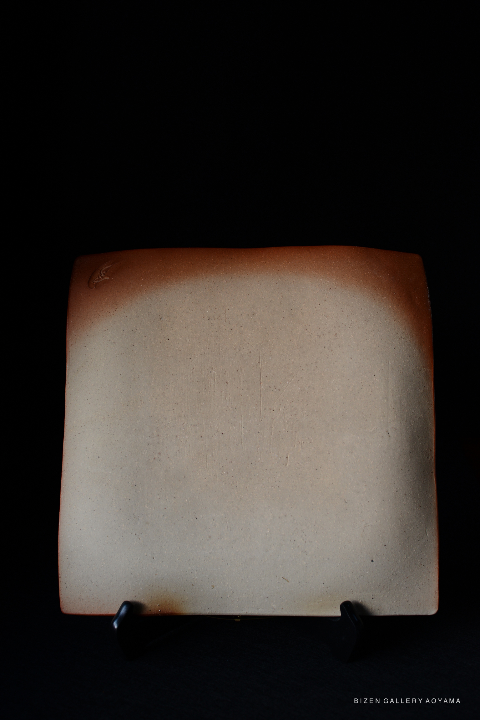 A minimalist beige ceramic plate displayed against a dark background. The plate features a smooth surface with subtle textures and a faint gradient of color towards the edges.