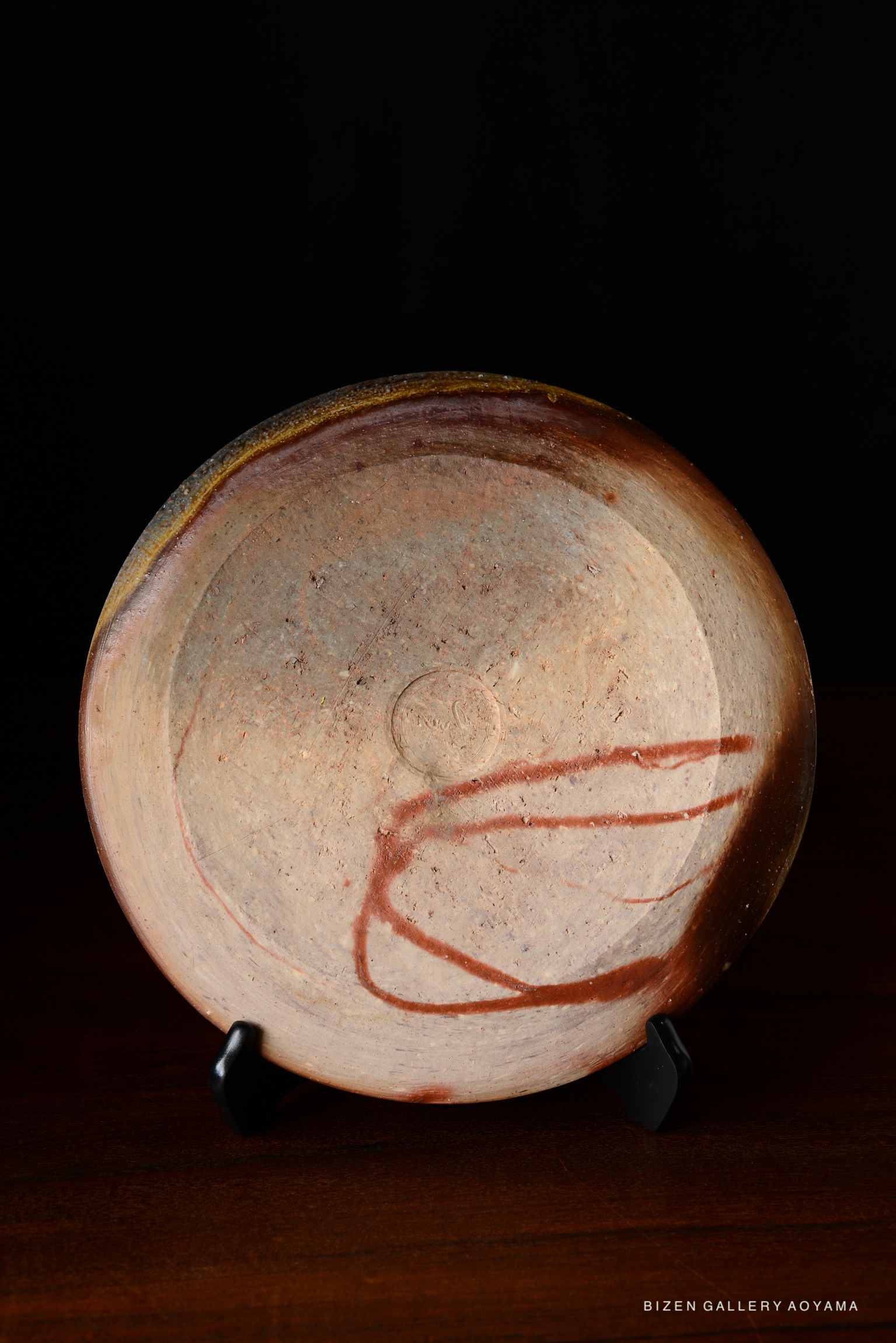 A ceramic plate with a smooth, natural surface displaying earthy tones and a subtle abstract design, showcased on a black stand.