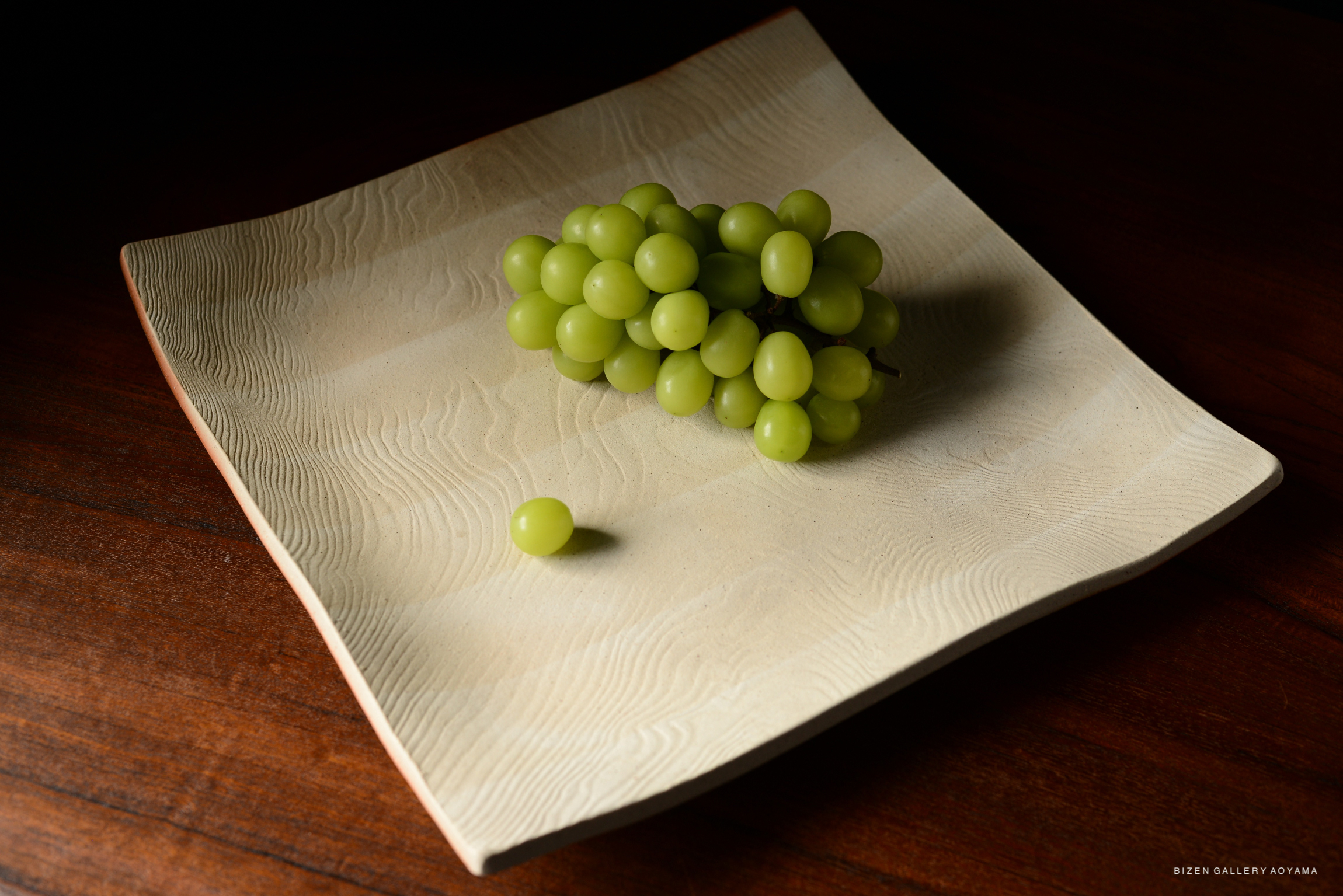 A rectangular ceramic plate with a textured surface, displaying a cluster of green grapes, set on a wooden table.