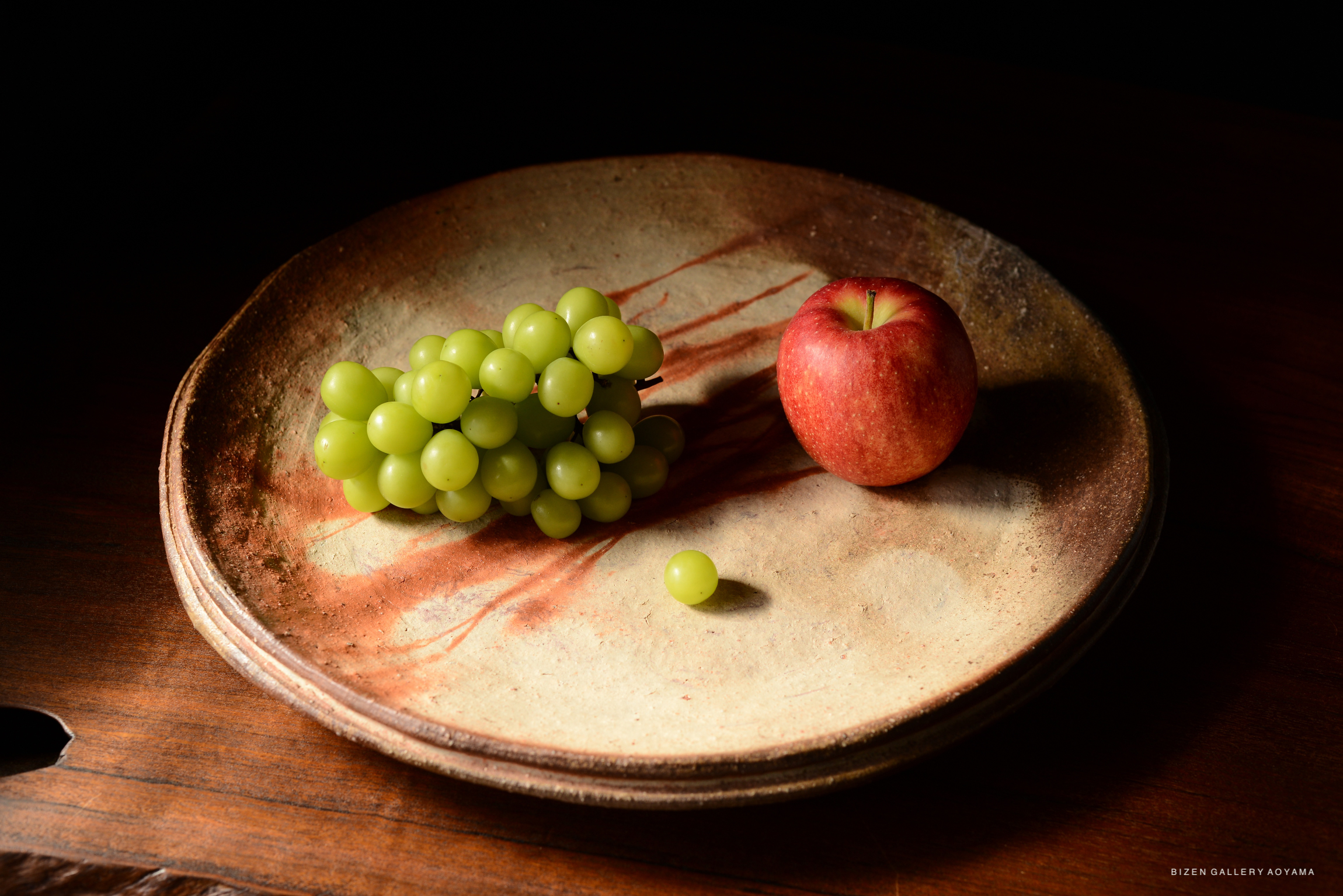 A still life composition featuring a cluster of green grapes and a red apple resting on a rustic ceramic plate with earthy tones.