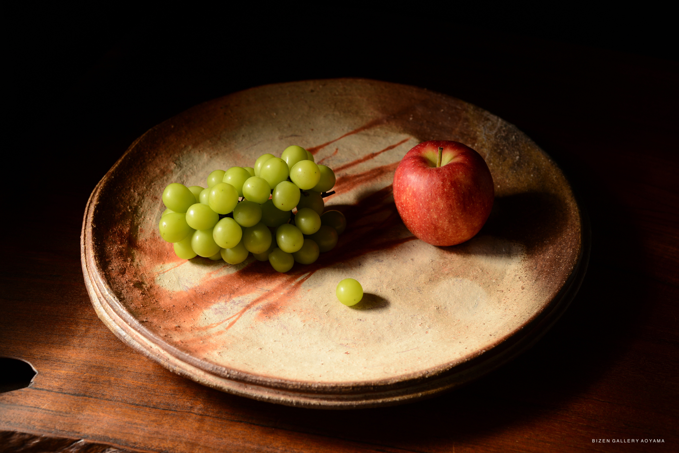 A still life featuring a cluster of green grapes and a red apple arranged on a textured ceramic plate.