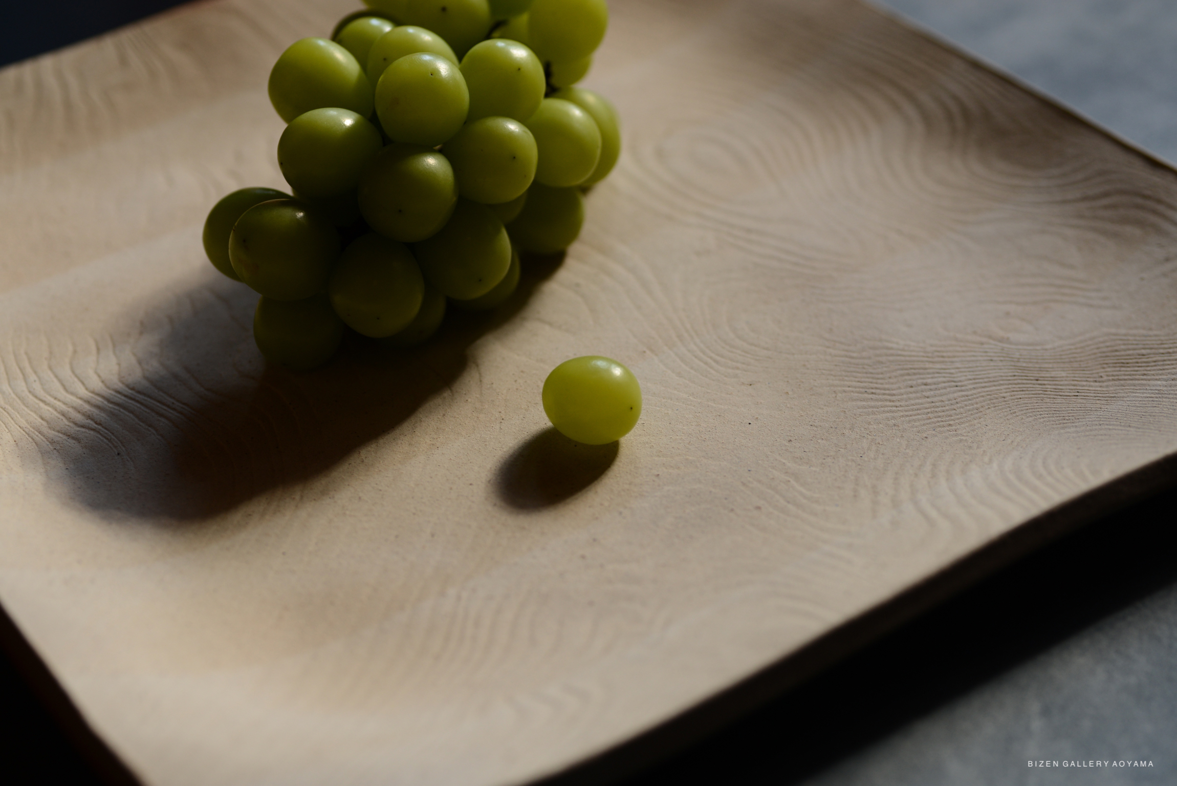 A close-up view of a cluster of green grapes placed on a textured, pale ceramic plate, with one grape rolled away from the cluster.