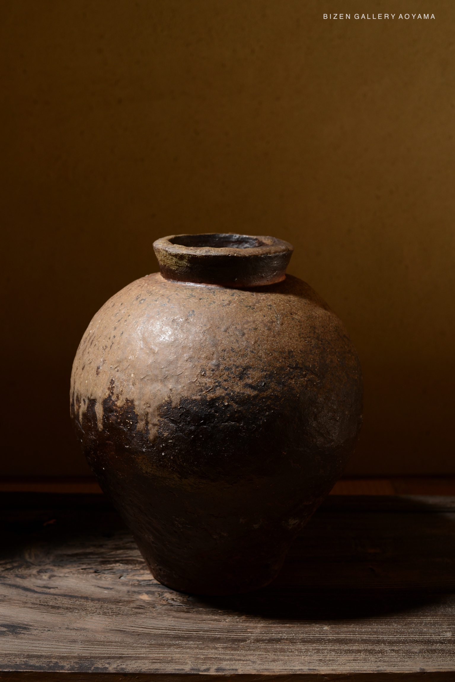 A traditional ceramic Tsubo vase with a rounded body and a narrow neck, featuring earthy tones and a textured surface, displayed on a wooden table against a plain background.
