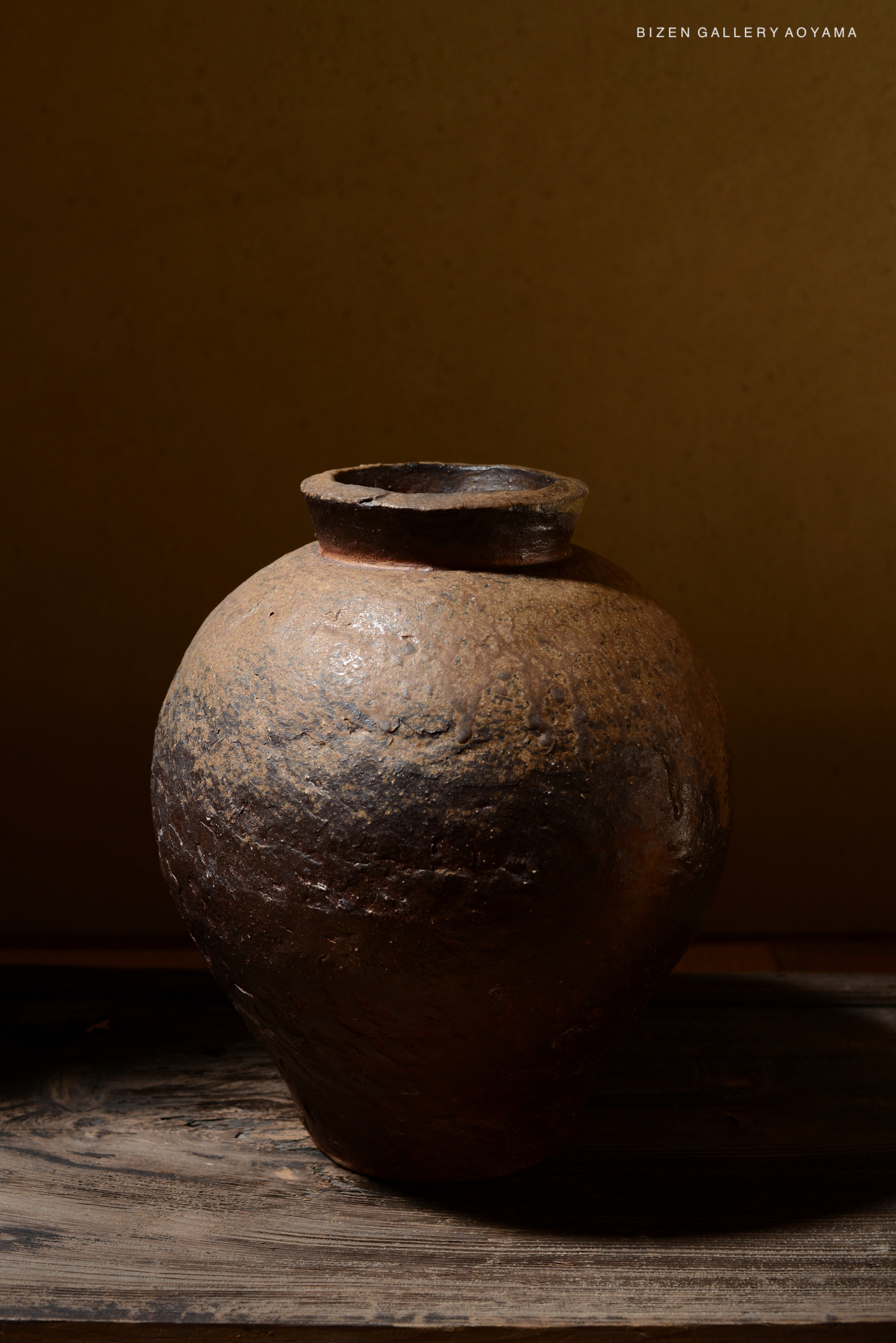 A rustic Bizen pottery tsubo (jar) with a rounded shape and earthy tones, placed on a wooden surface and set against a muted background.