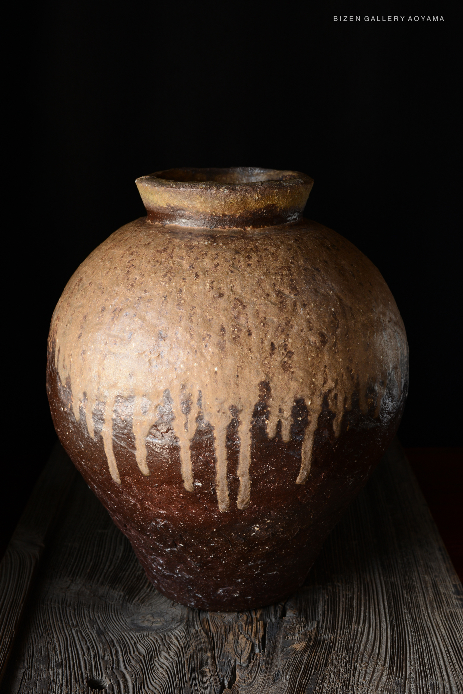 A large, rustic tsubo (Japanese storage jar) with a textured surface and dripped glaze, displayed on a wooden surface against a dark background.