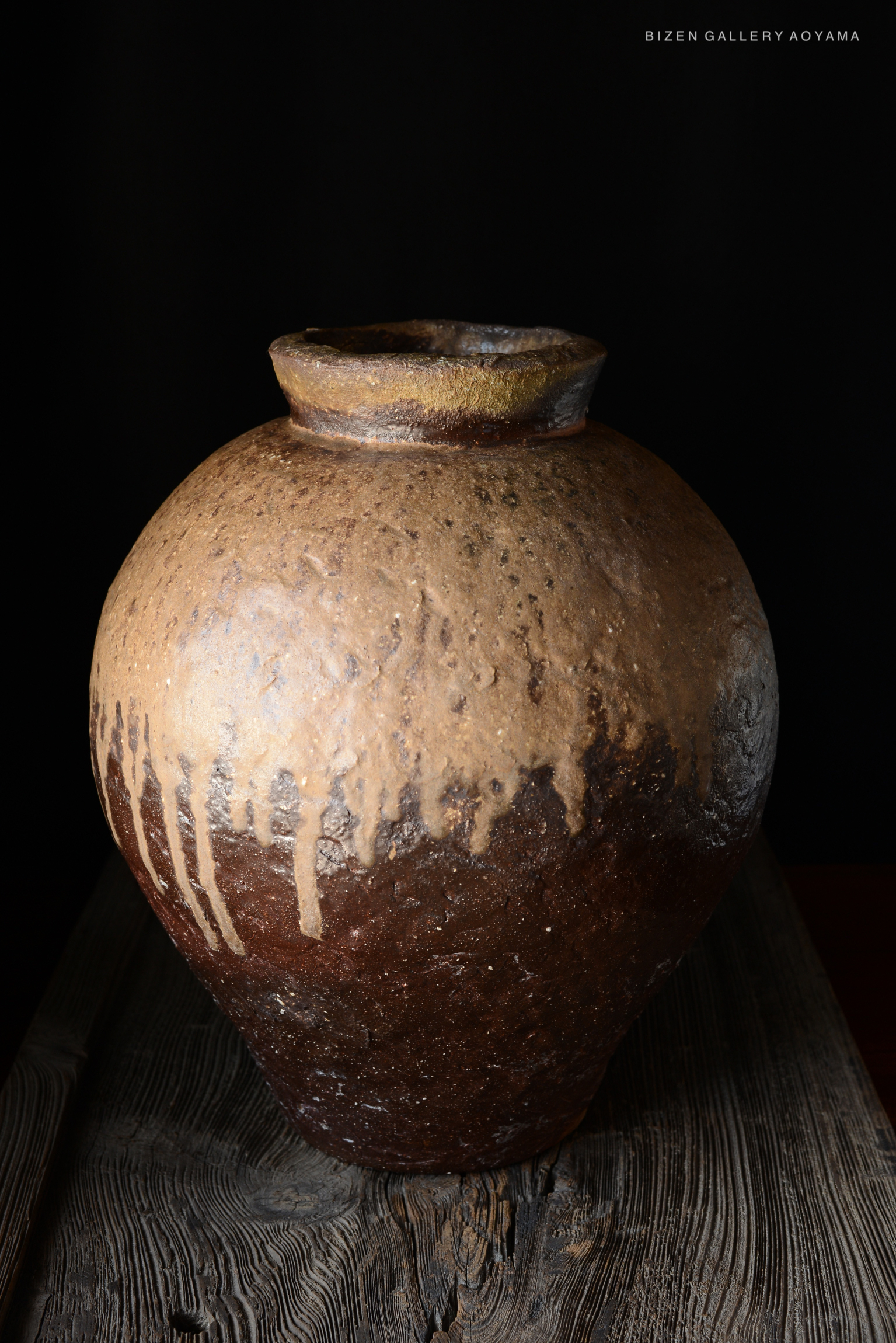 A large, rustic ceramic tsubo (jar) with a textured surface and a dark color palette, displaying drips of glaze, set on a wooden table against a black background.