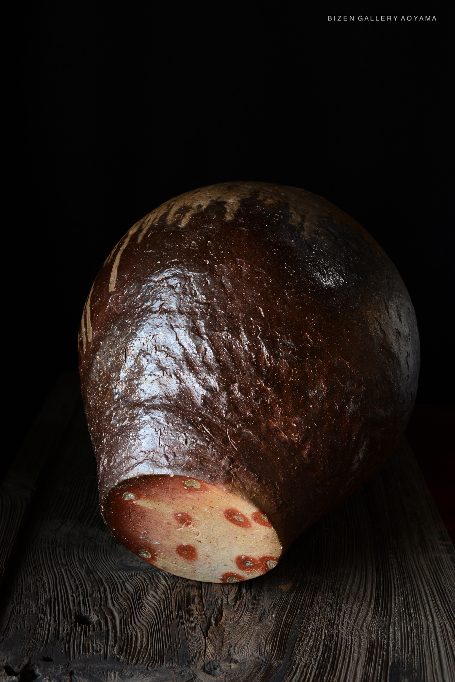 A large, rounded ceramic tsubo with a textured, dark brown exterior and a light-colored base showing unique patterns, set against a black background.