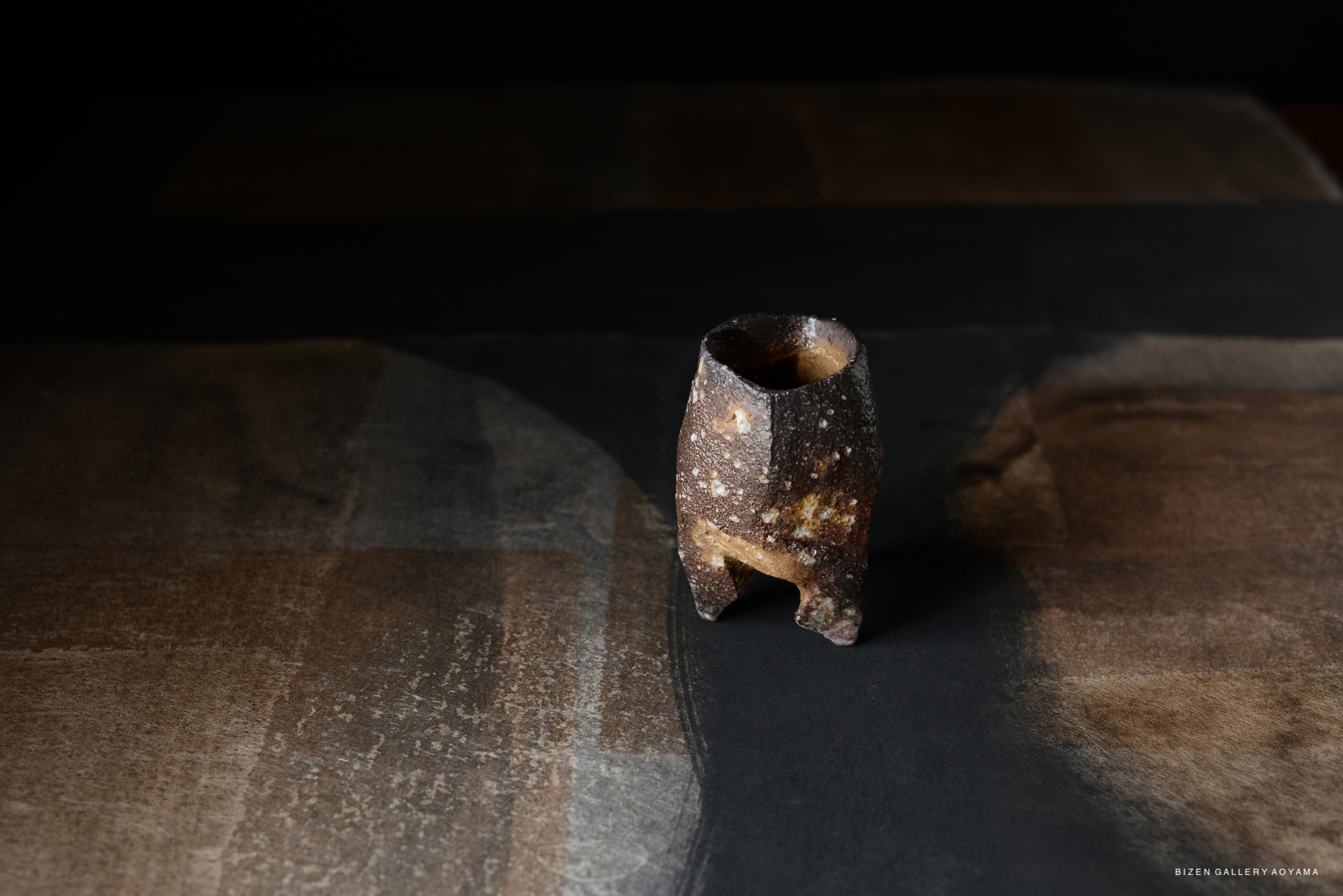 A small, rustic ceramic vessel with a textured surface, placed on a dark, abstract background.
