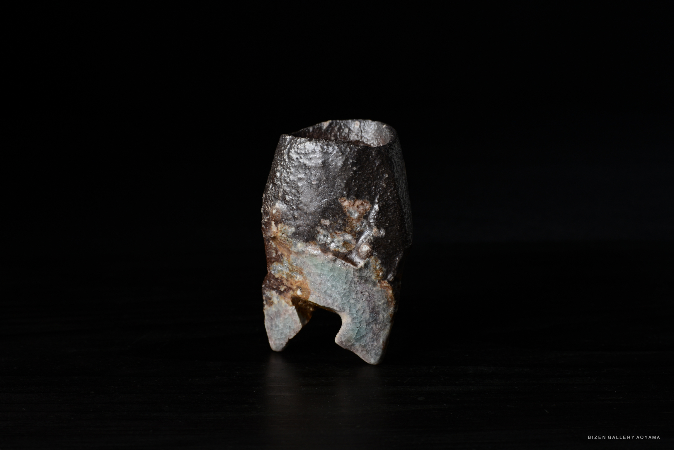 A close-up view of a unique, textured ceramic piece with a dark, rough exterior and subtle color variations, set against a black background.