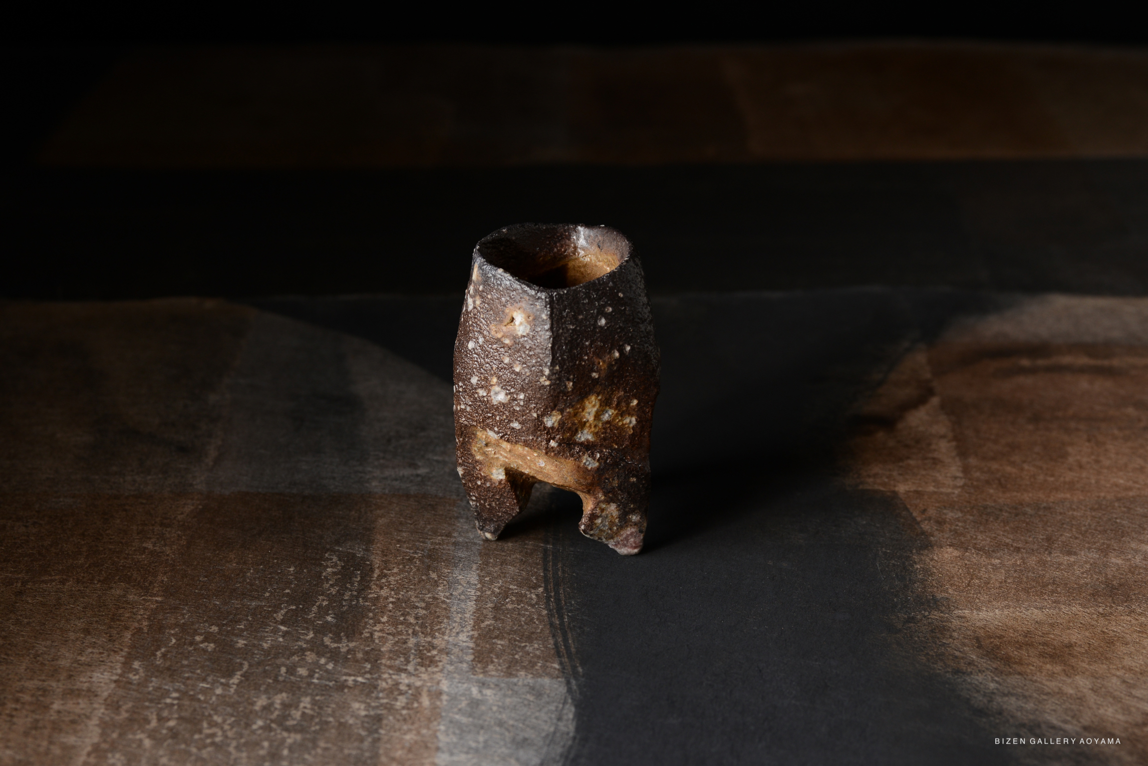 A textured, dark ceramic vase with a unique shape, placed on a dark, patterned surface.