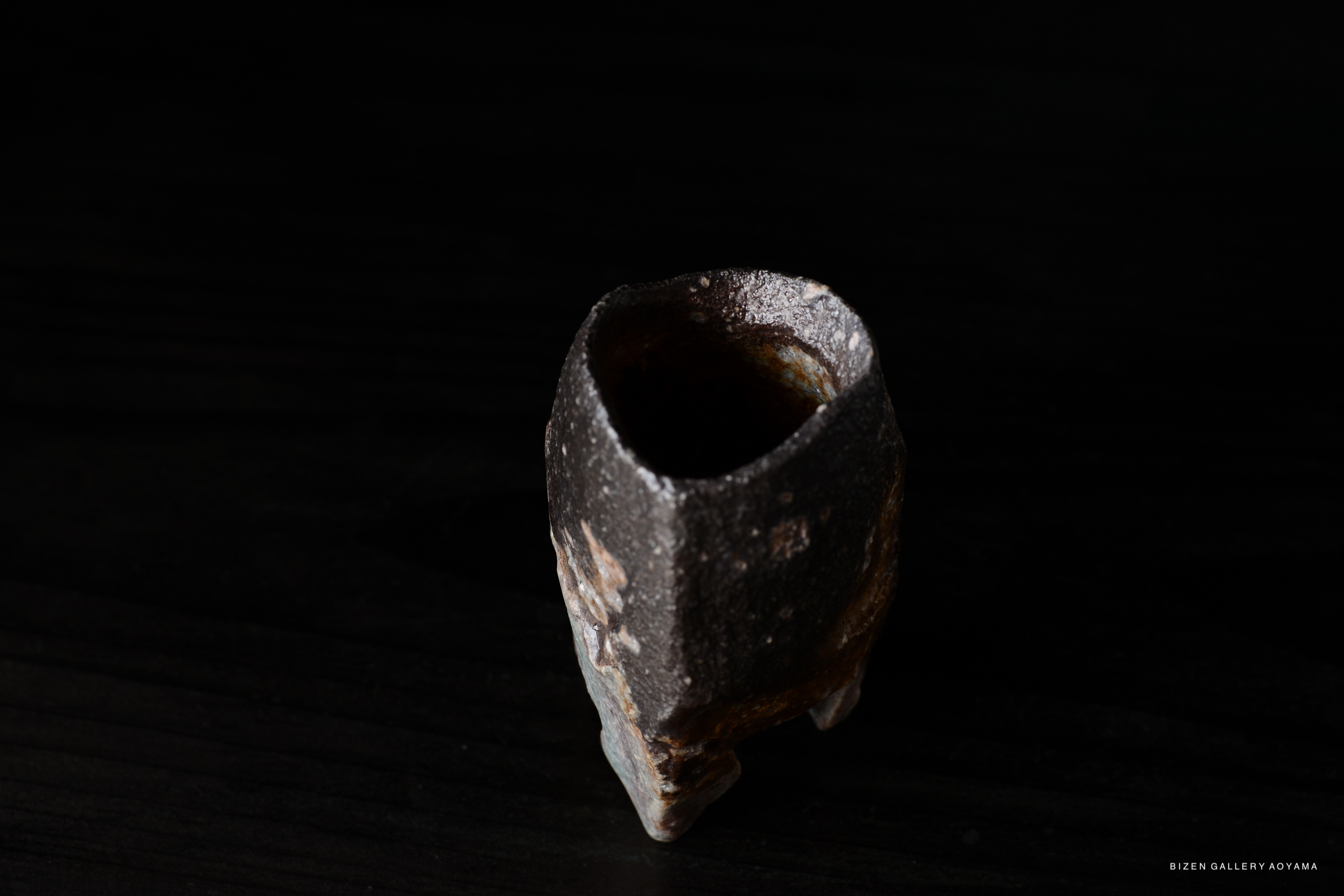 A close-up of a textured ceramic vessel with an irregular shape, featuring dark colors and a slightly glossy interior, placed on a black surface.
