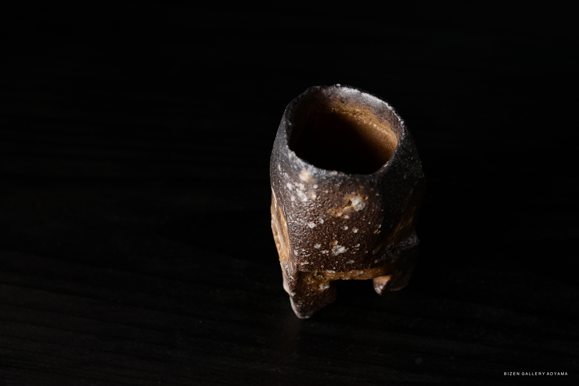 A close-up view of an artistic ceramic vessel with a textured surface and organic shape, set against a dark background.