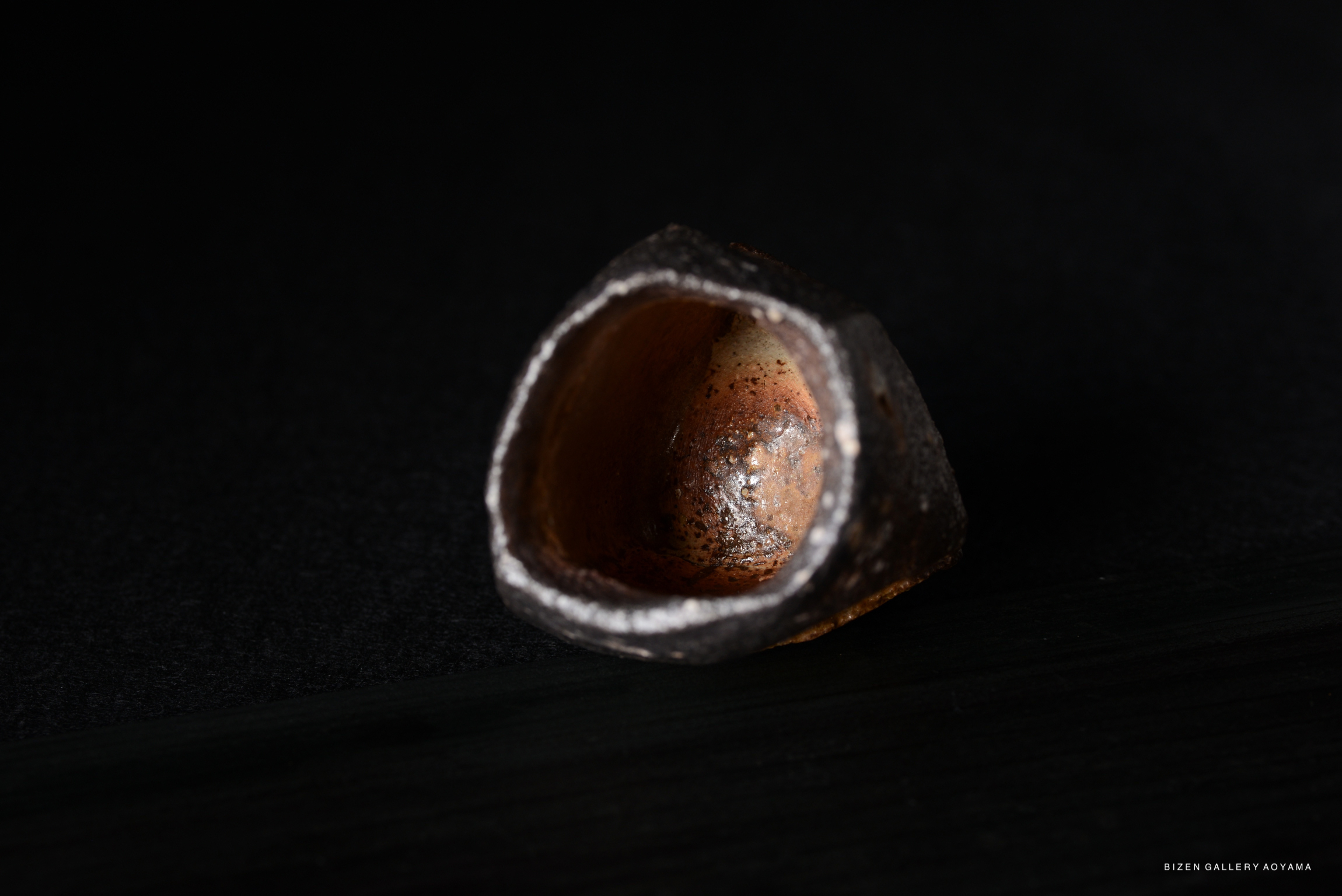 Close-up of a small, irregularly shaped ceramic vessel with a textured exterior and a smooth, warm-toned interior, resting on a dark surface.