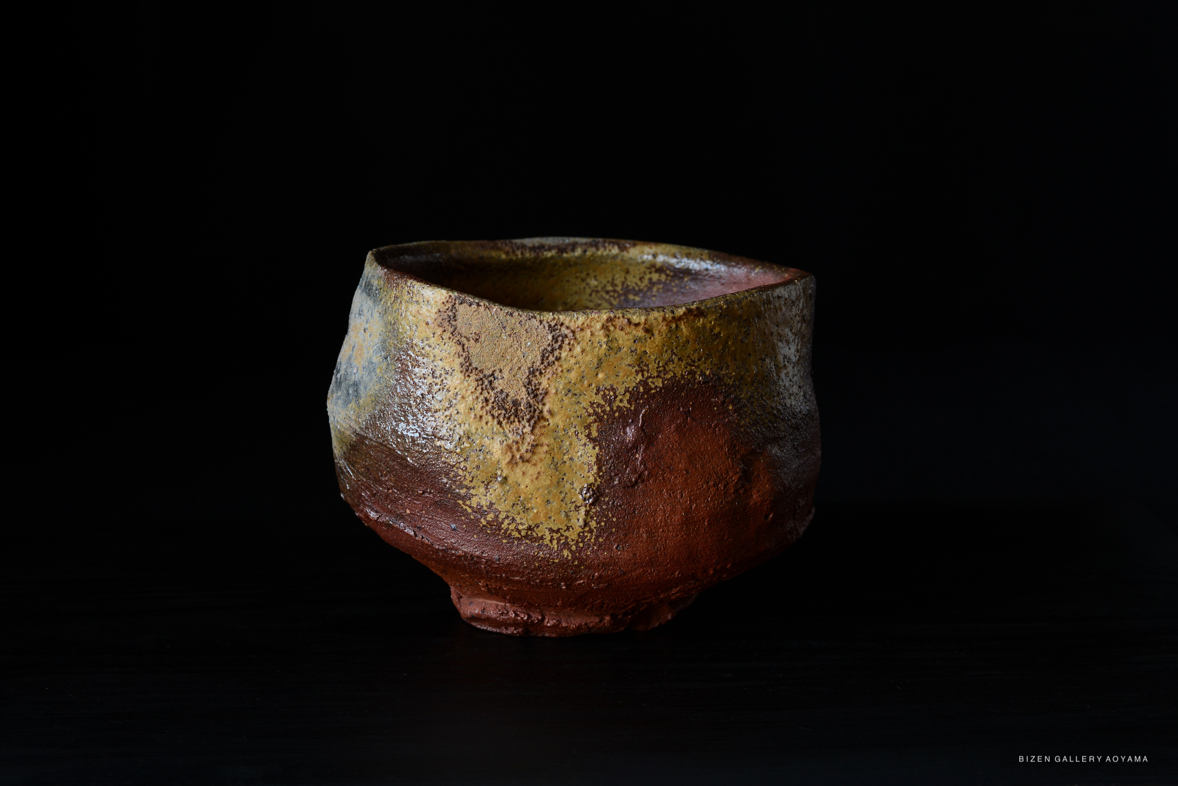 A rustic Bizen tea bowl with textured surface in earthy tones of yellow, brown, and orange, displayed against a black background.