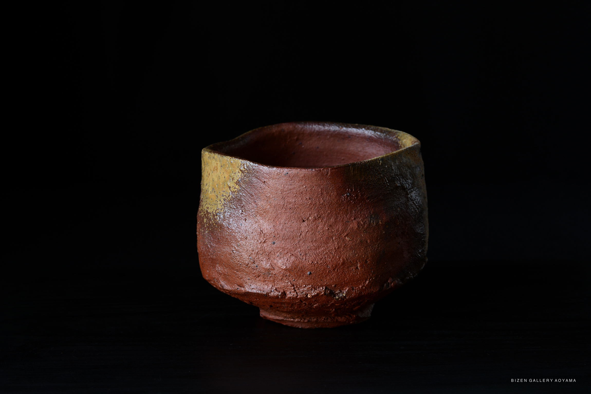A rustic Bizen tea bowl with a reddish-brown texture and a slightly uneven rim, set against a black background.