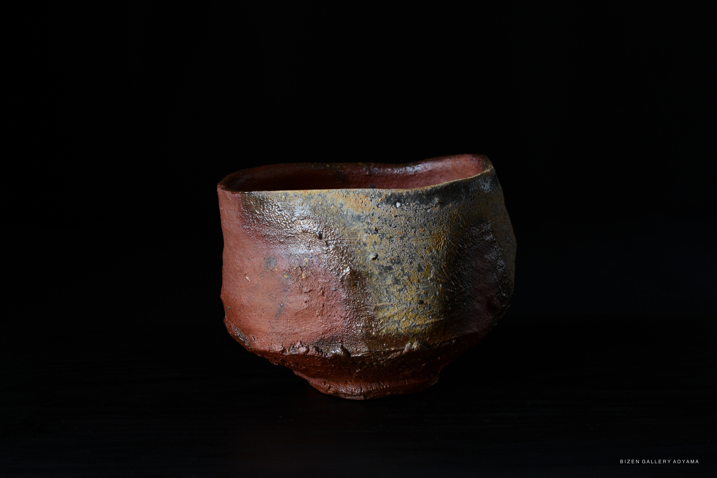 A handcrafted Bizen tea bowl with an irregular shape and a mix of earthy colors, displayed against a dark background.