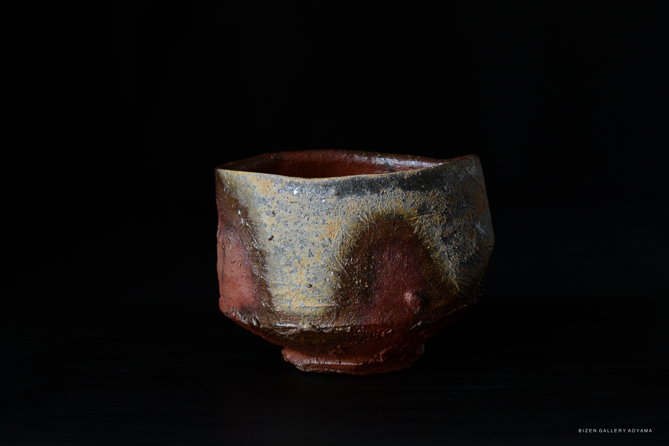 A traditional Bizen tea bowl with a unique shape and earthy tones, featuring a textured surface and a dark background.