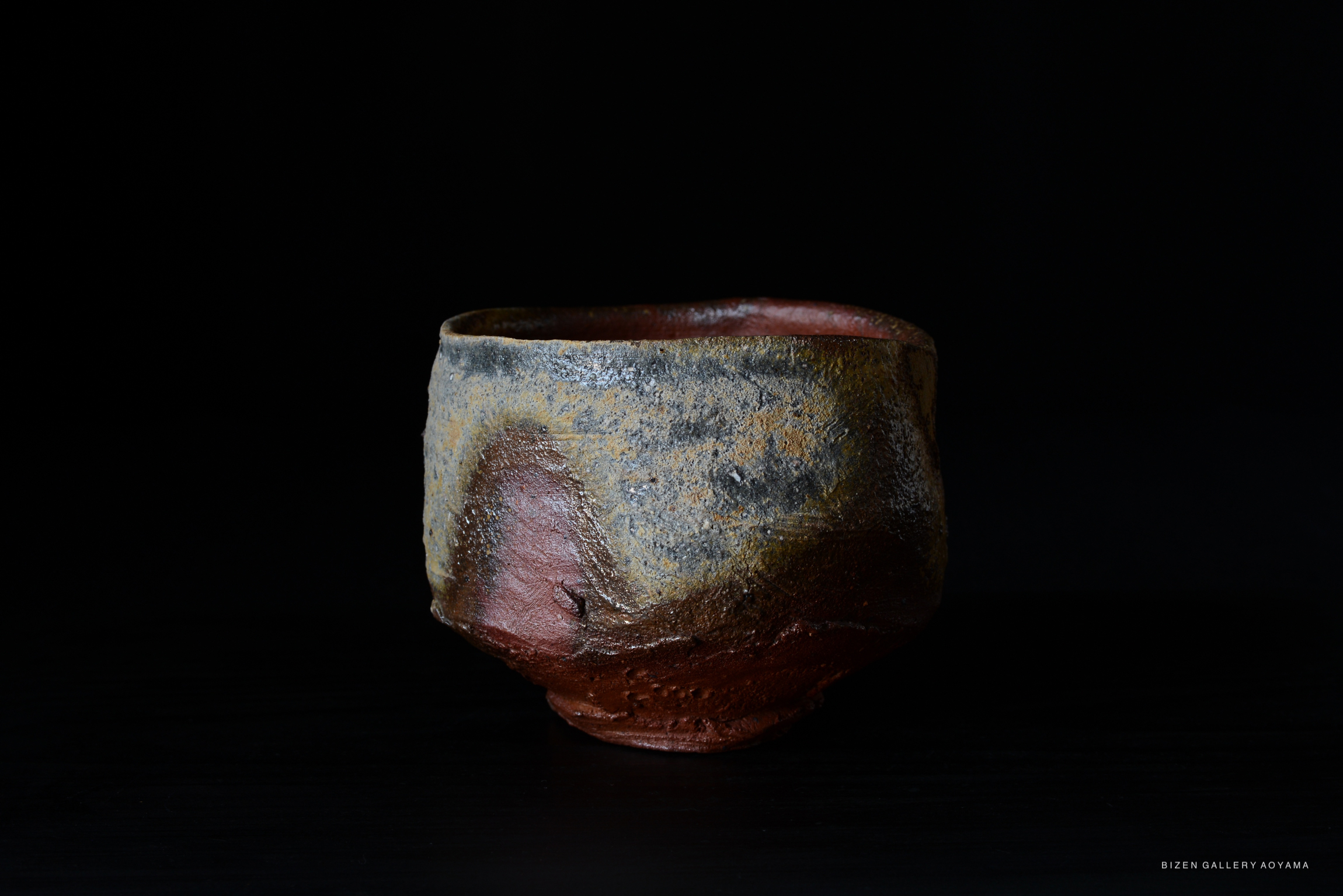 A handcrafted Bizen tea bowl with a rustic finish, featuring earthy tones and unique textures, displayed against a black background.