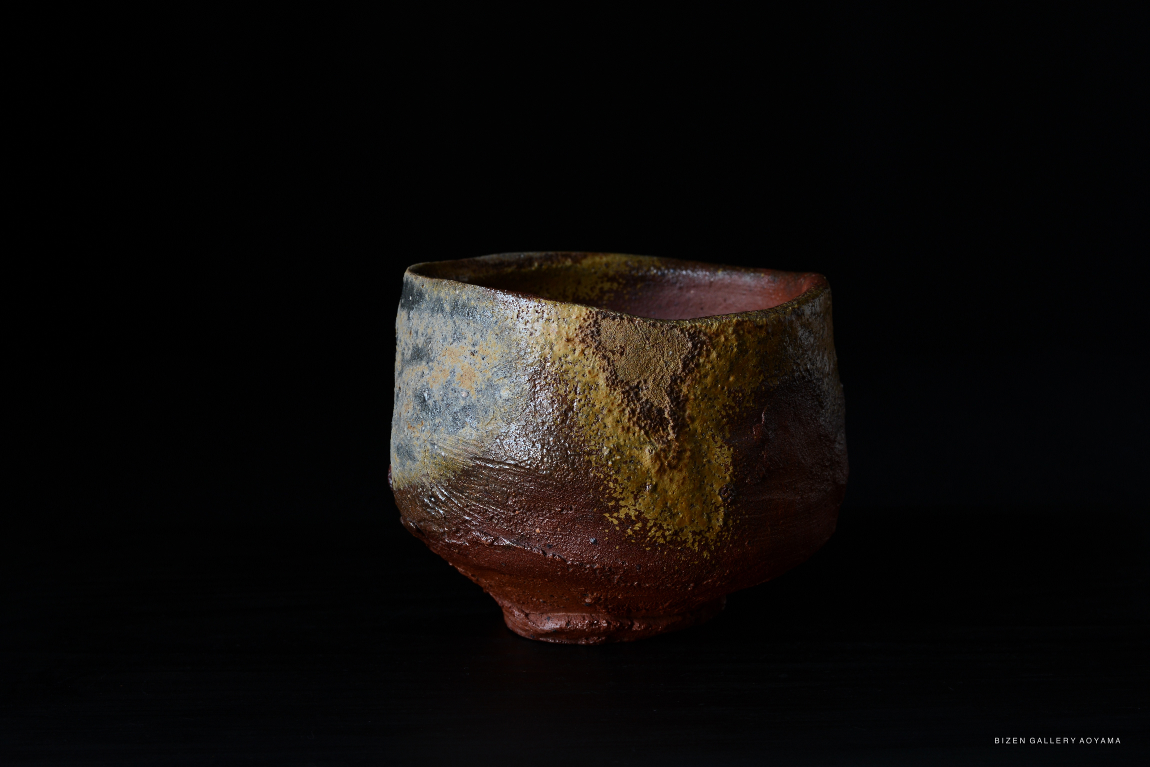 A handmade Bizen pottery tea bowl featuring a textured surface with earthy tones of brown and gray, displayed against a black background.