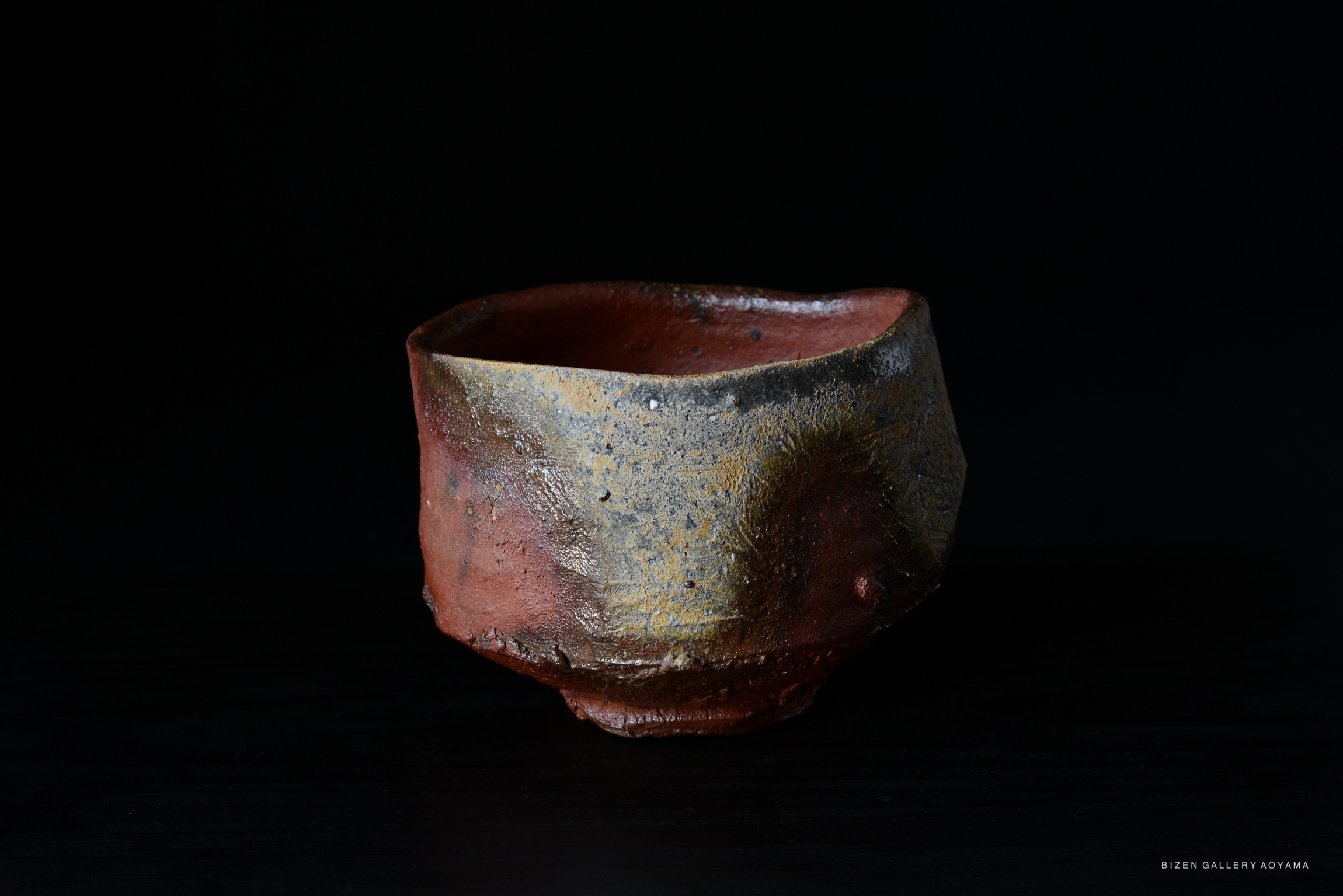 A rustic Bizen tea bowl with an uneven, textured surface featuring earthy tones of red and gray, set against a black background.