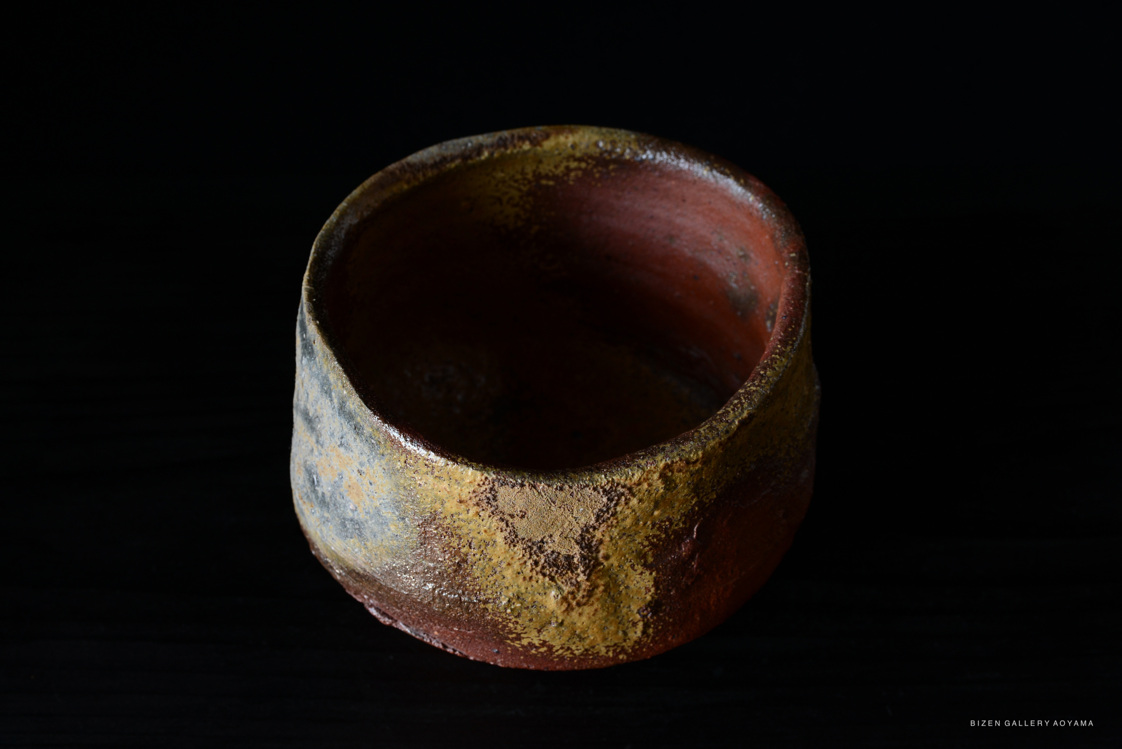 A rustic Bizen tea bowl with a textured surface showcasing earthy colors, set against a dark background.