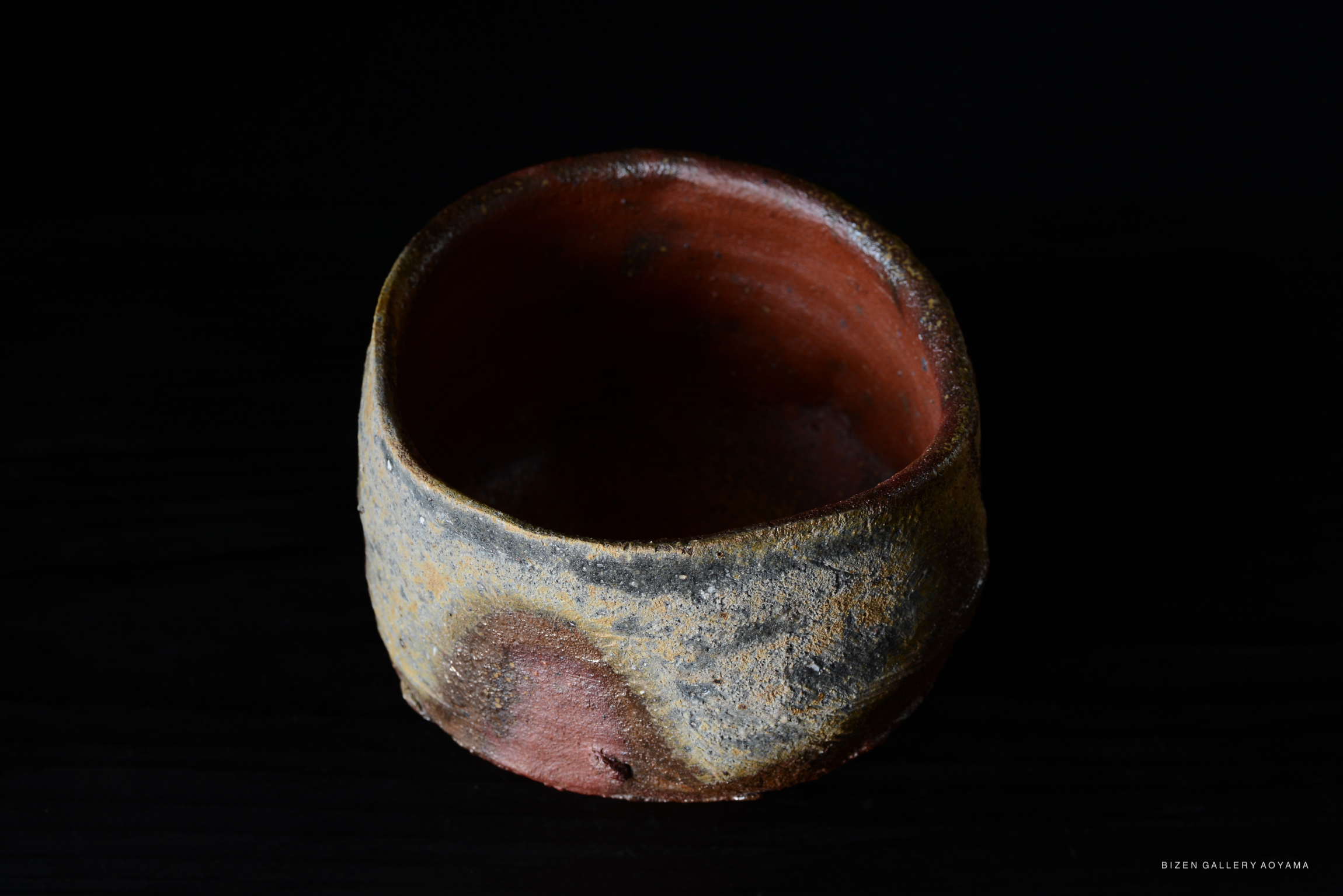 A top view of a Bizen tea bowl showcasing its rustic, earthy texture and rich colors. The bowl has a subtle gradient with shades of brown, orange, and gray, set against a dark background.