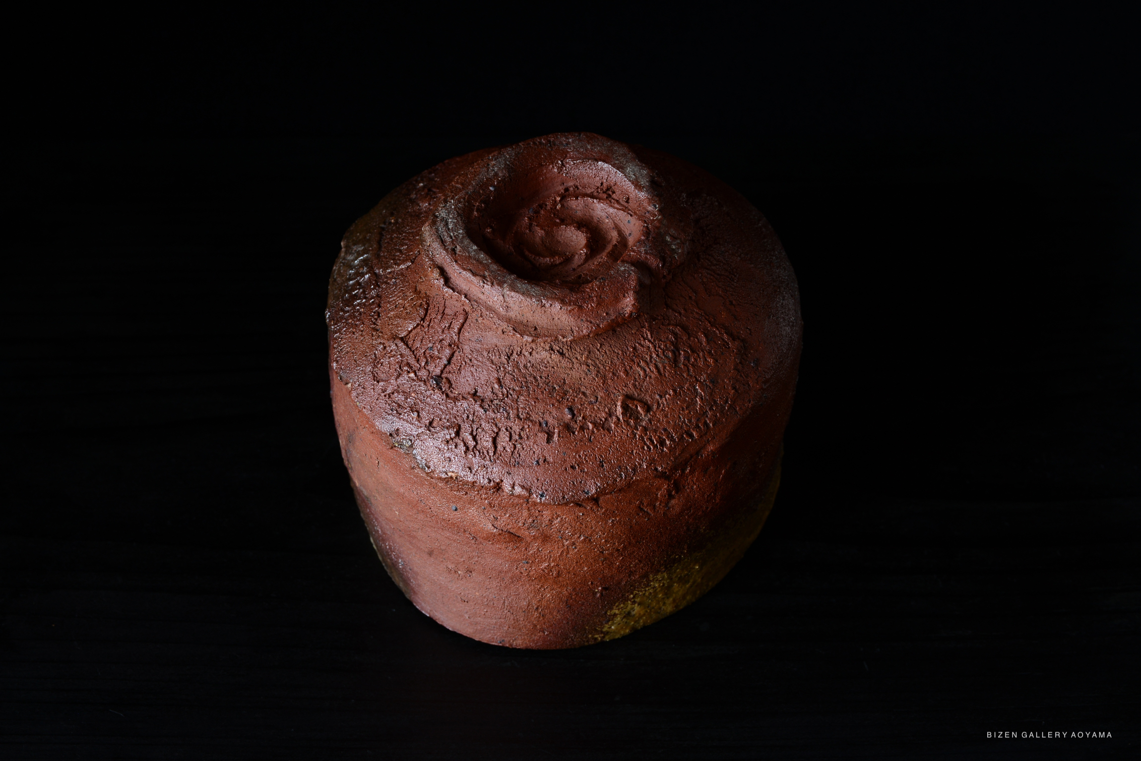 A Bizen tea bowl with a textured, reddish-brown surface, featuring a spiraled top, displayed against a dark background.