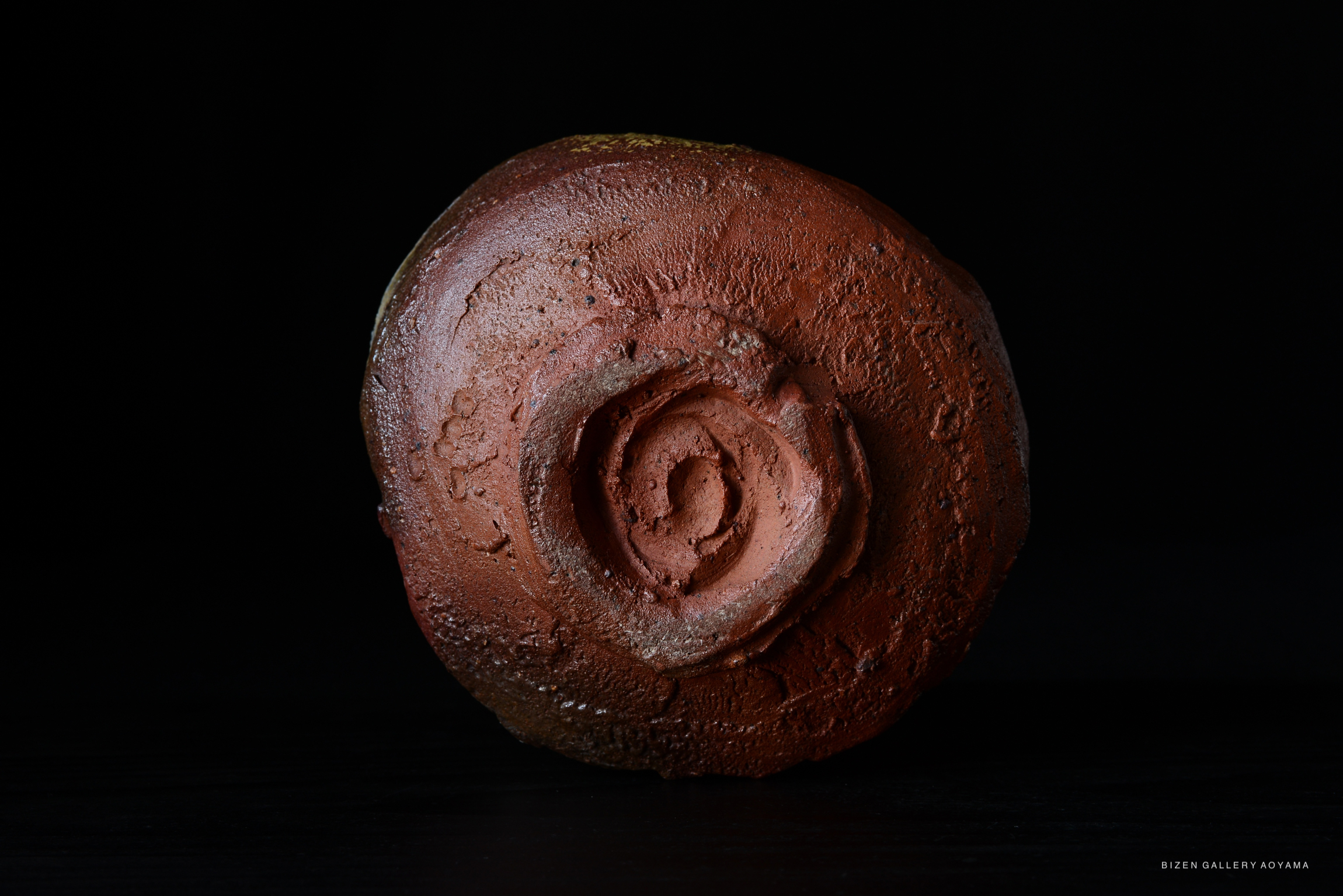 A close-up view of a Bizen tea bowl with a textured, earthy surface and a spiral design on the base, set against a black background.