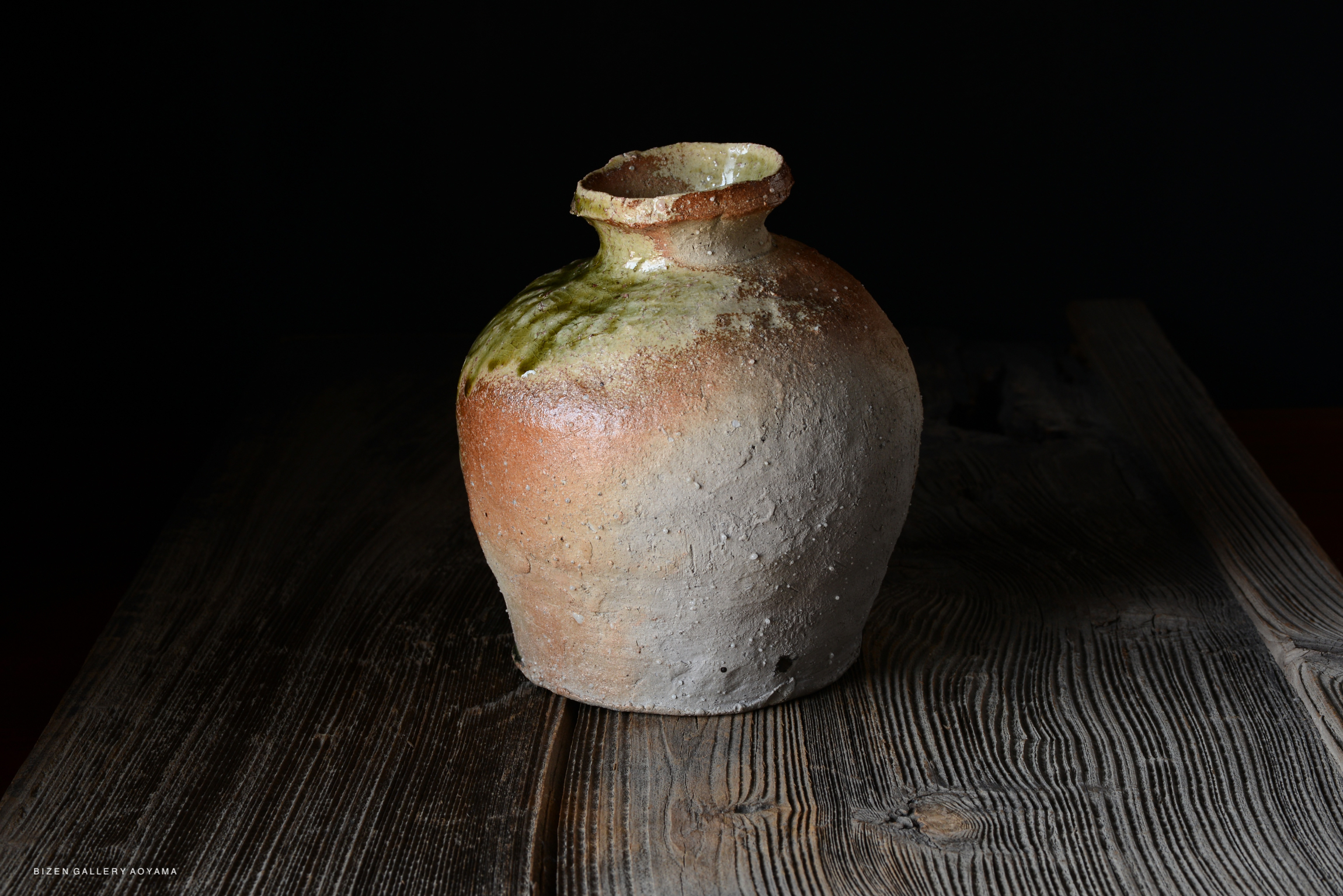 A rustic ceramic vase with a rounded shape, featuring a mix of earthy tones and a glossy green glaze at the top, positioned on a wooden surface.