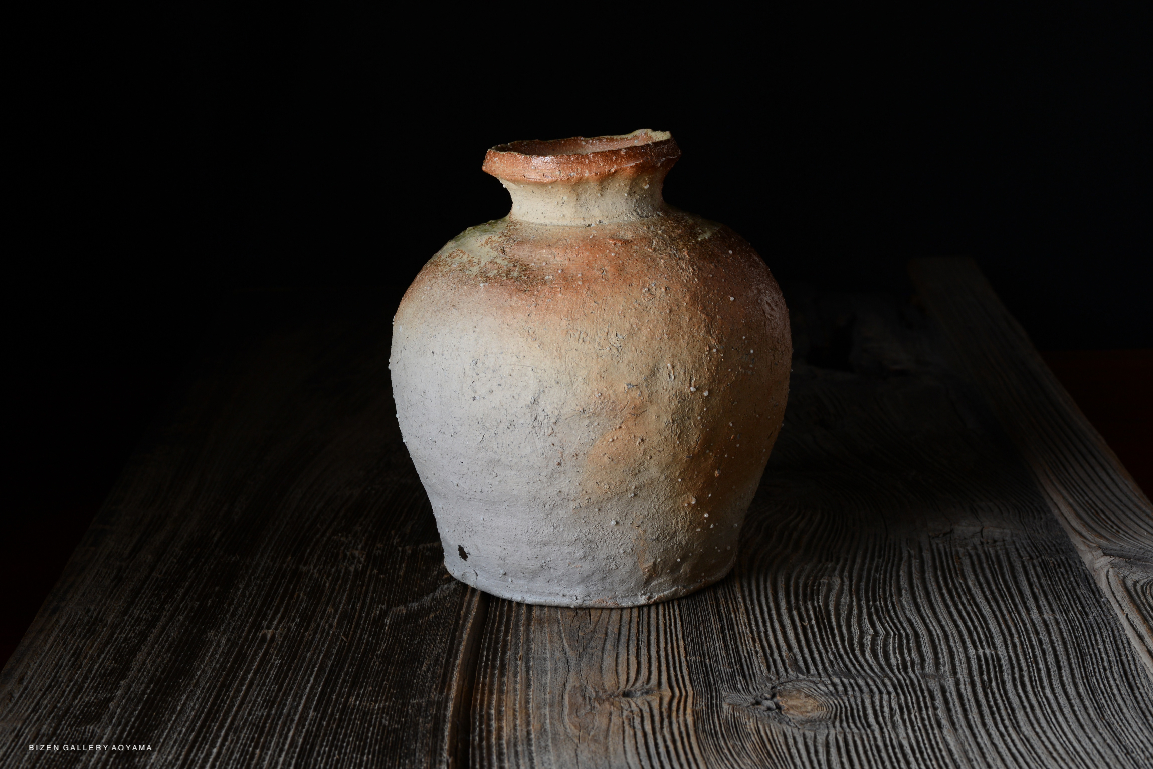 A traditional ceramic pot with a rounded body and a narrow neck, featuring a textured surface with earthy tones, sitting on a wooden table against a dark background.