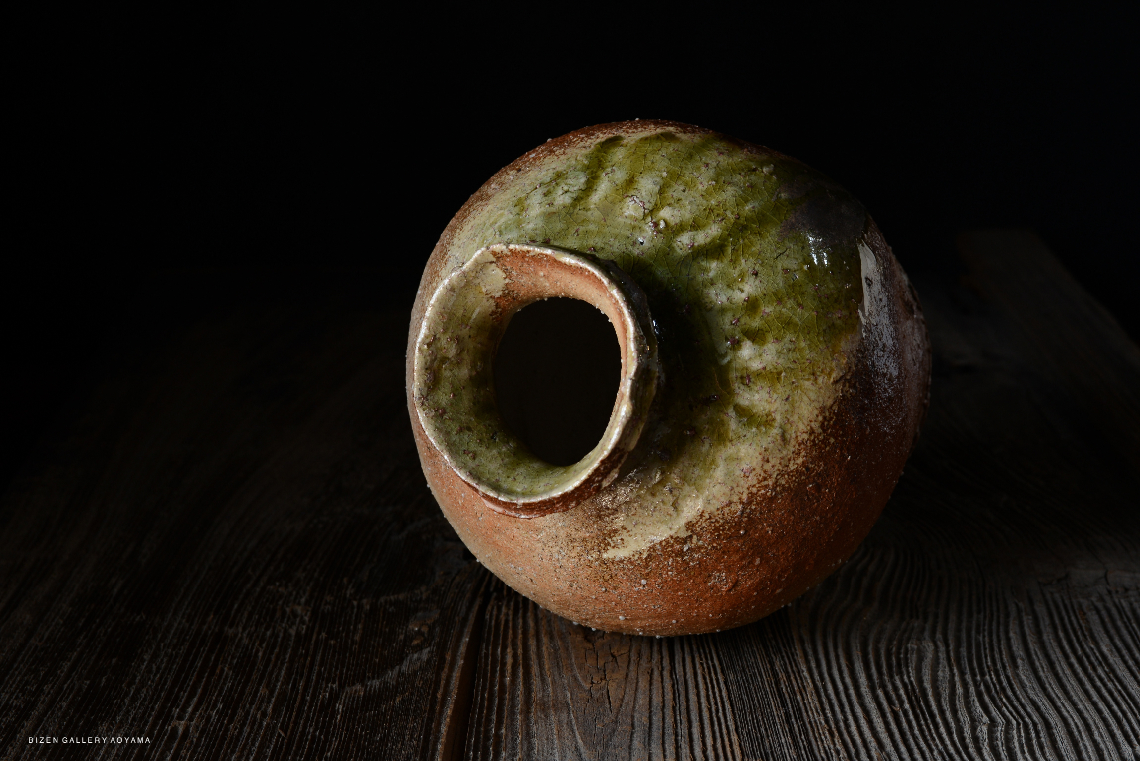 A rustic ceramic pot with a green and brown glaze, featuring a narrow neck and a round body, placed on a wooden surface with a dark background.