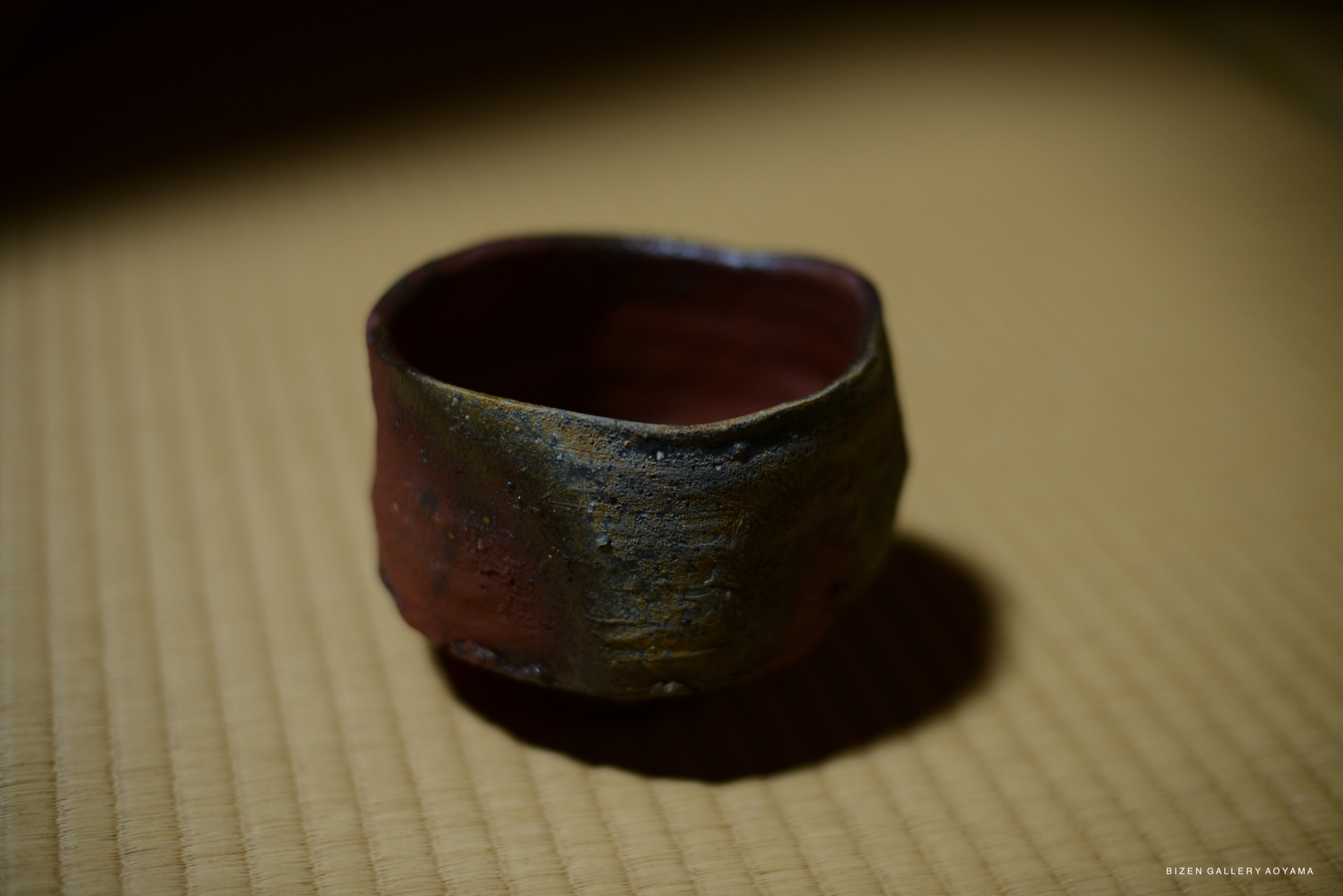 A close-up view of a Bizen tea bowl with a rustic design, featuring dark and reddish tones on a tatami mat background.