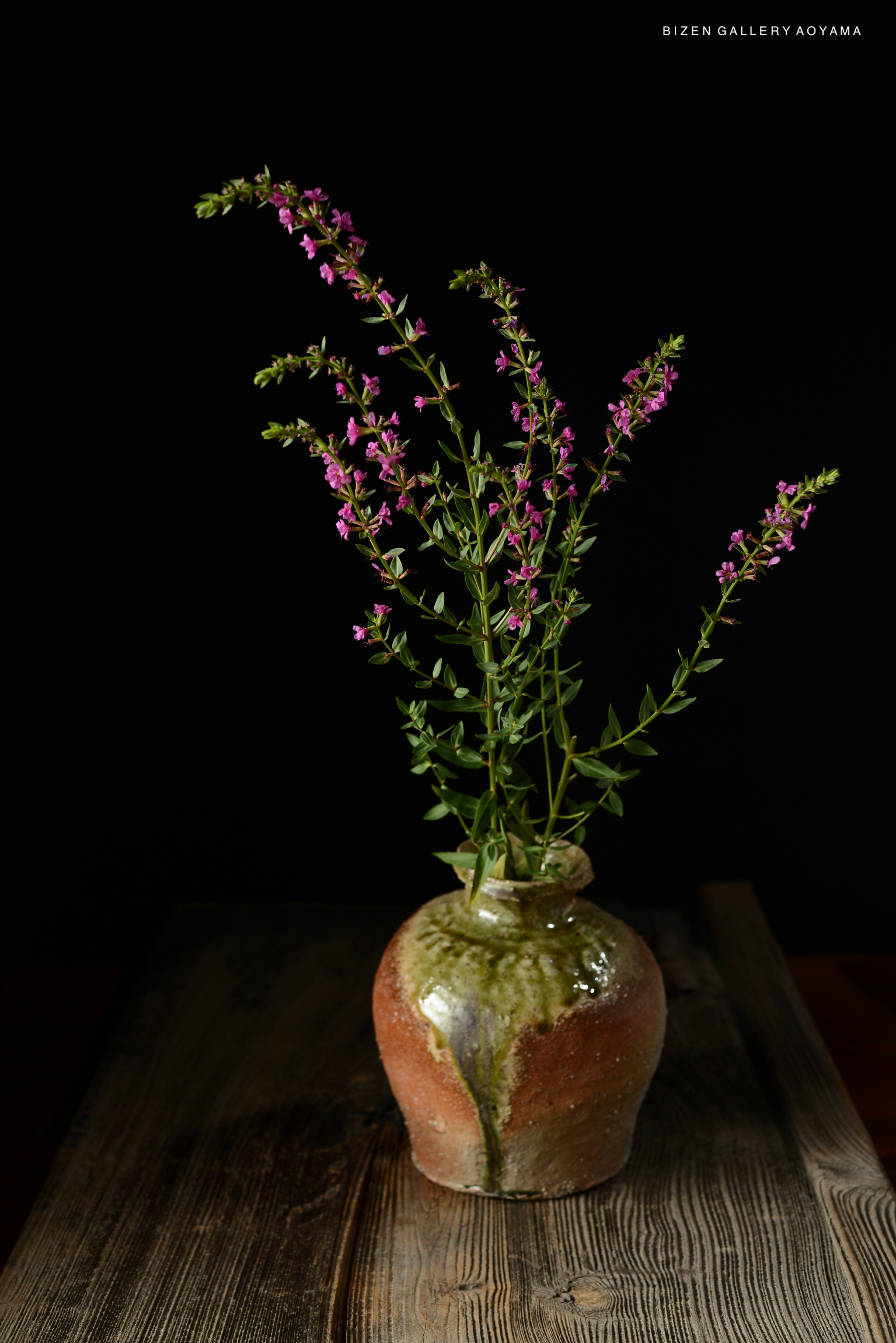 A ceramic vase with a muted earthy color and a glossy green glaze on top, holding slender green stems with small pink flowers, set against a black background.