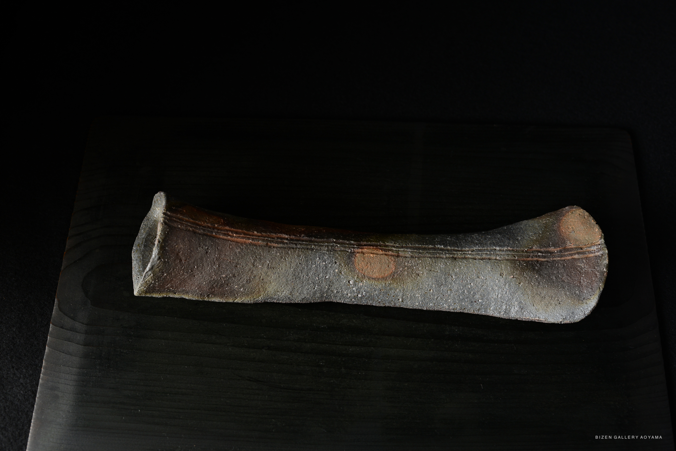A dark, textured ceramic piece resembling a long, narrow dish, set against a black background.