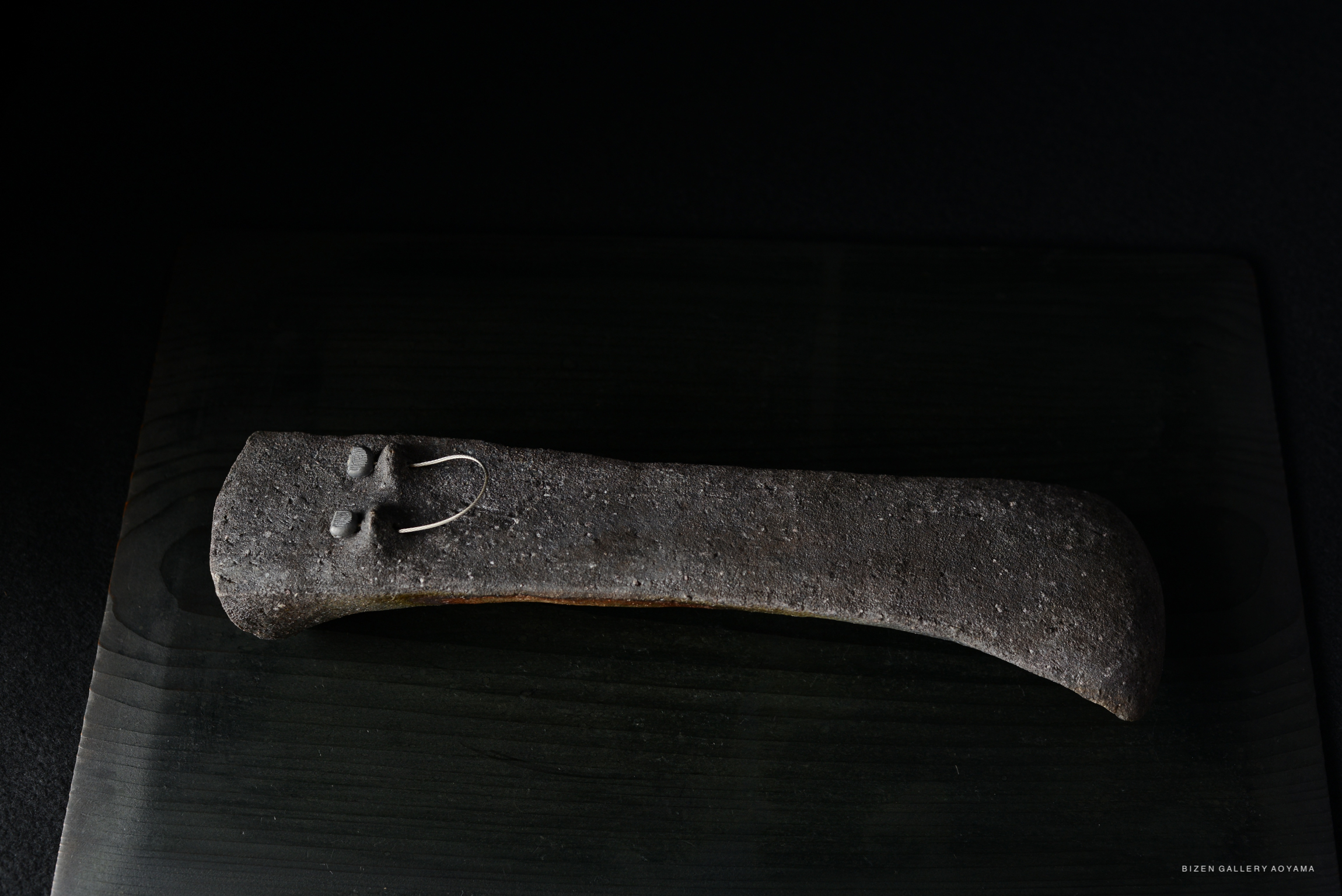 Ceramic object with a textured surface, featuring small circular details and a metallic hook, positioned on a dark background.