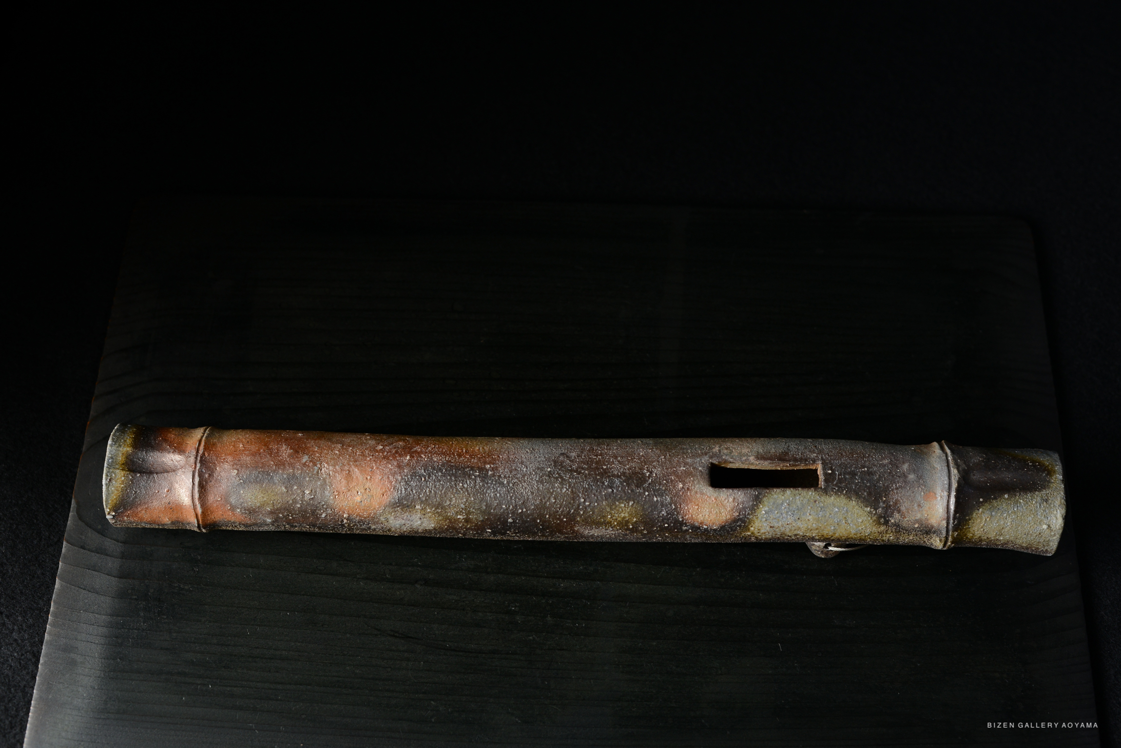 A close-up view of a traditional Bizen pottery flute, showcasing its earthy colors and textured surface, placed on a dark wooden background.